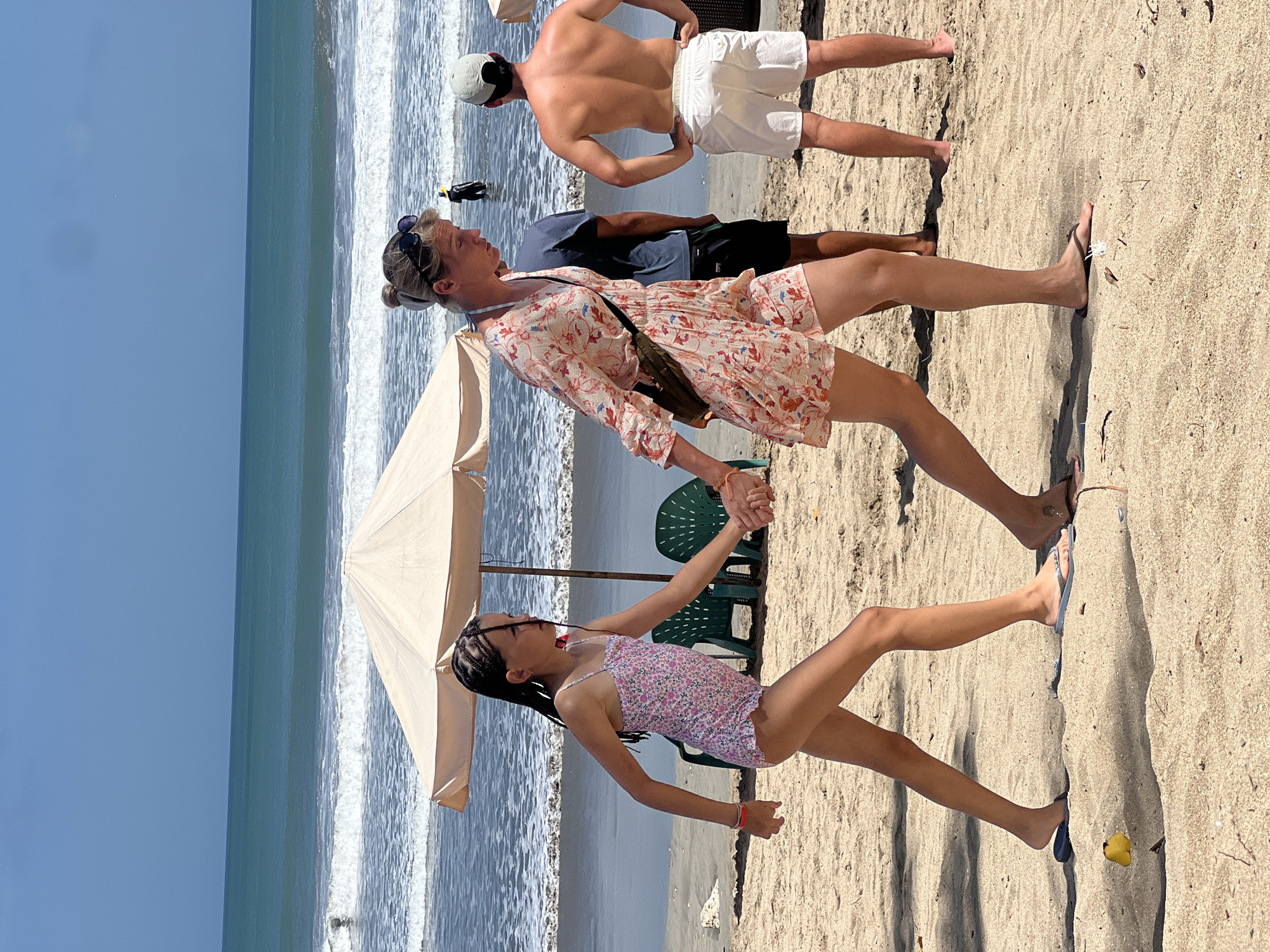 In Kuta, Indonesia, on a sunny day at the beach, a woman and a young girl walk hand-in-hand on the sand. The woman wears a floral-print dress and a crossbody bag. The girl is in a floral swimsuit. They are walking away from the ocean, which has gentle waves breaking on the shore. In the background, two men stand on the beach. One man, wearing white shorts, faces away from the camera, while another man in a dark shirt stands between him and the woman and girl. Beach chairs and an umbrella are visible near the water's edge. The sand is light brown and appears dry.