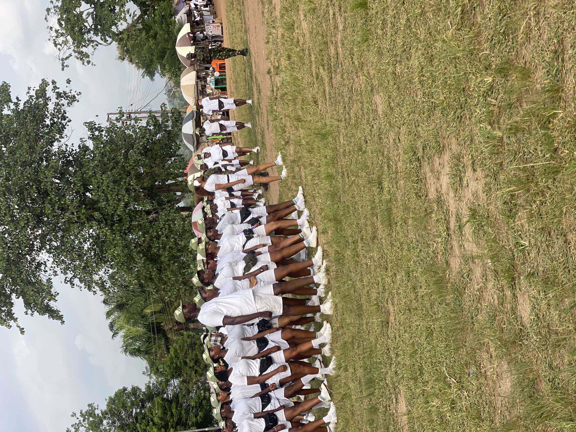 A group of young people, appearing to be participants in a program or event, stand in formation on a grassy field in Emure-Ekiti, Nigeria. They are dressed in matching white t-shirts, white shorts, and green caps. Many are also wearing black fanny packs around their waists and white socks with white sneakers. The group is arranged in rows, facing to the right of the frame, with some individuals looking forward and others to the side.
In the background, there are several market stalls with umbrellas providing shade. Behind the stalls, there are trees, utility poles, and electrical wires, indicating an outdoor, semi-rural or peri-urban setting. A soldier in camouflage uniform is visible walking to the right of the frame, past the market stalls. The ground is covered with short, dry grass and some dirt paths. The sky is overcast.