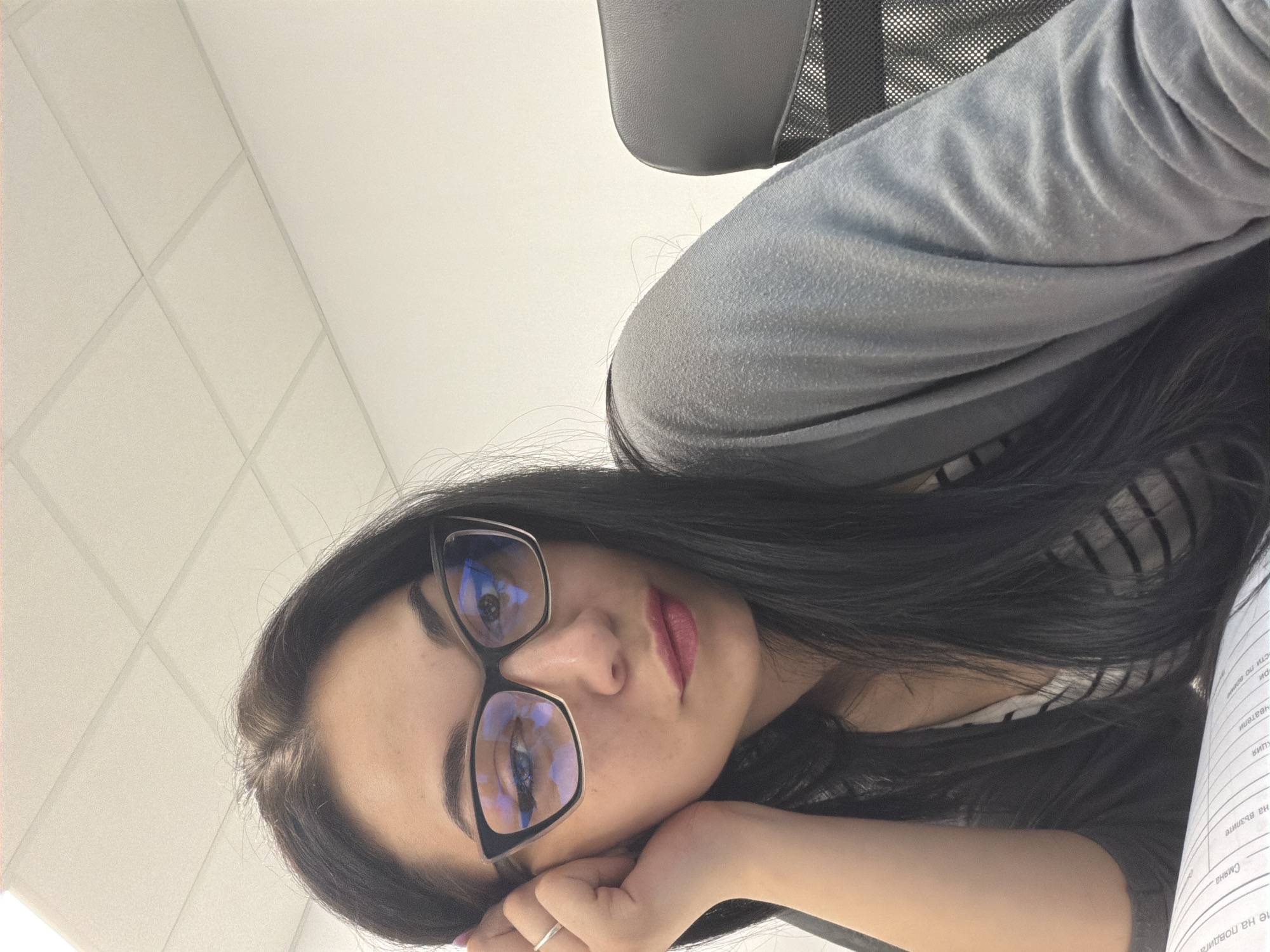 A young woman with long, dark hair and glasses is leaning on her hand, looking at the camera. She is wearing a gray cardigan over a striped shirt. The background appears to be an office environment with a white wall and a drop ceiling. The image was taken in Stara Zagora, Bulgaria.
