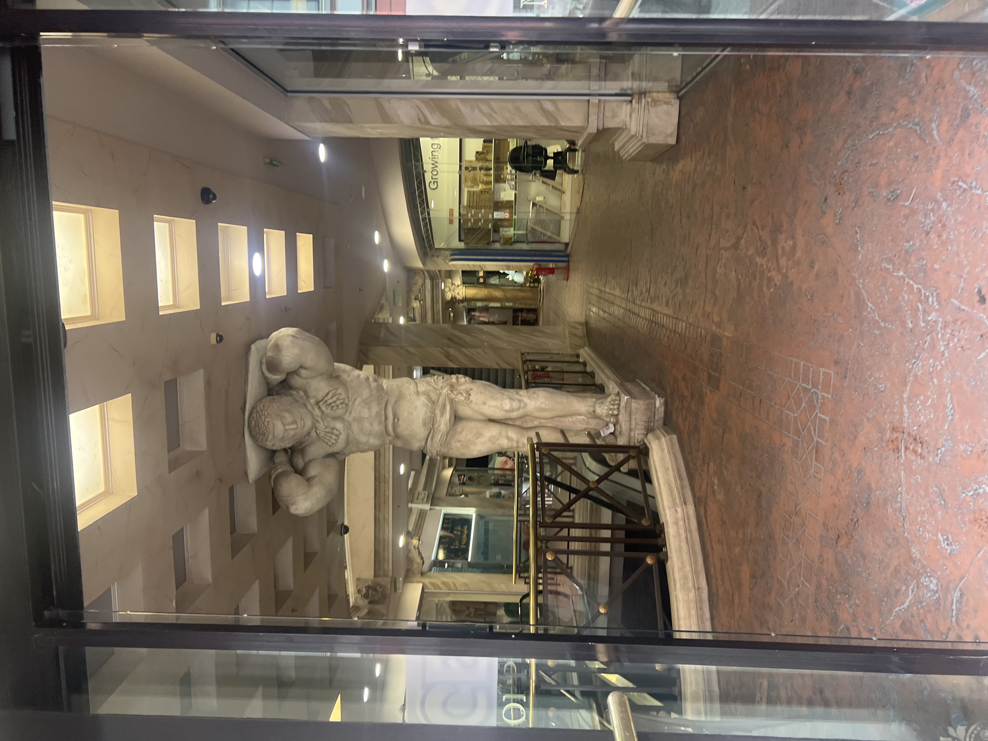 The image depicts the interior of a building in Plovdiv, Bulgaria, viewed through a glass door or window. The main focus is a large, reclining statue of a muscular male figure, likely made of marble or a similar light-colored stone. The statue is positioned on a slightly elevated platform with a metal railing. The surrounding area is decorated in a classical style, featuring marble columns, textured walls with recessed lighting, and an ornate ceiling. A hallway stretches into the distance, lined with shops and architectural details. The floor is a patterned brick or tile in reddish-brown hues. A storefront with a sign that reads "Growing" is visible further down the hallway. The lighting is a mixture of natural and artificial, creating a warm and inviting atmosphere.