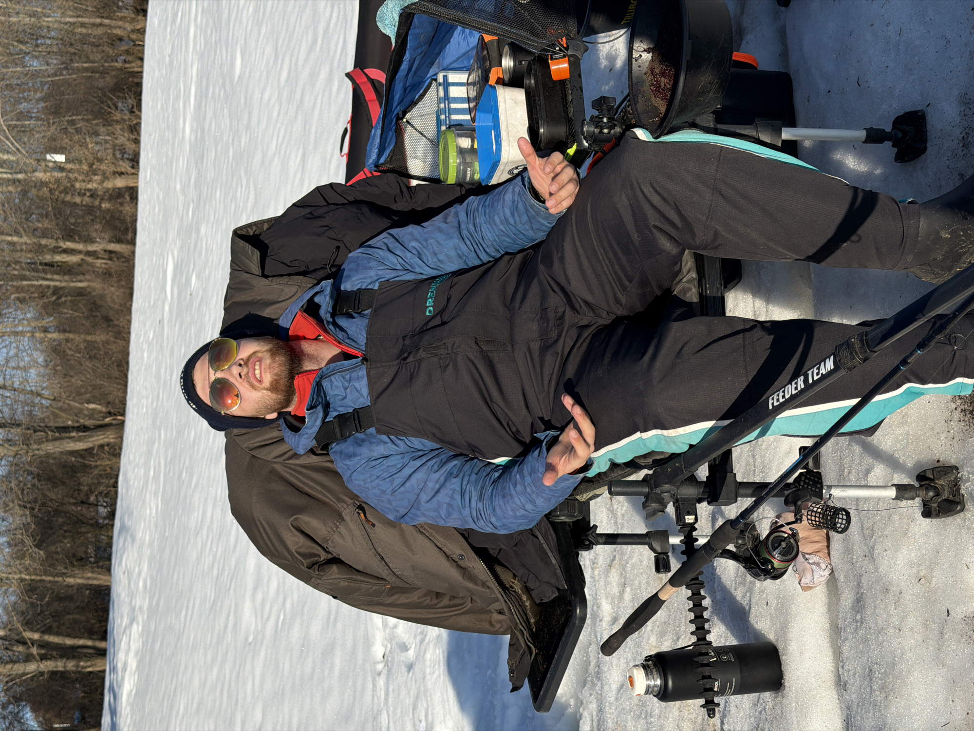A man wearing sunglasses, a beanie, a blue jacket, and black overalls with a turquoise stripe down the leg is sitting in a fishing chair on the snow. The chair is equipped with fishing rod holders, and a fishing rod is in one of the holders. There is a black bucket with fishing gear behind the chair. The background shows a snow-covered field and trees, typical of a cold climate. The location is likely in the vicinity of Chaikovsky, Russia, given the provided information. The man appears to be gesturing with his hands and smiling.