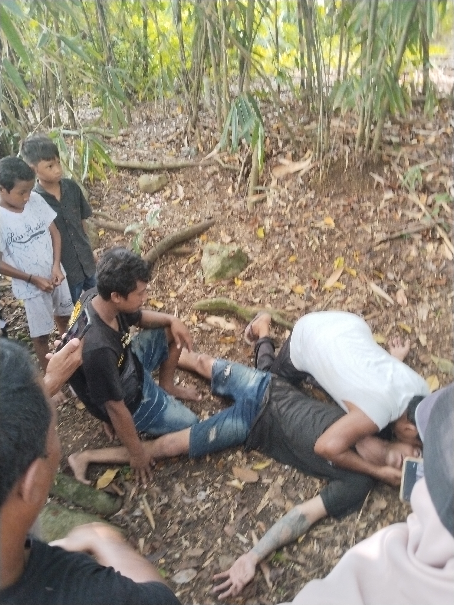 The scene is located outdoors in what appears to be a forested area in Pereng, Indonesia. The ground is covered in leaves and twigs with bamboo trees visible in the background. A man in a black shirt and blue jeans is lying on the ground with another man in a white shirt kneeling beside him, appearing to administer first aid. A man is taking a picture of the scene with their phone. Nearby, another man is kneeling and looking at the man on the ground. Two young boys are standing and looking at the scene.