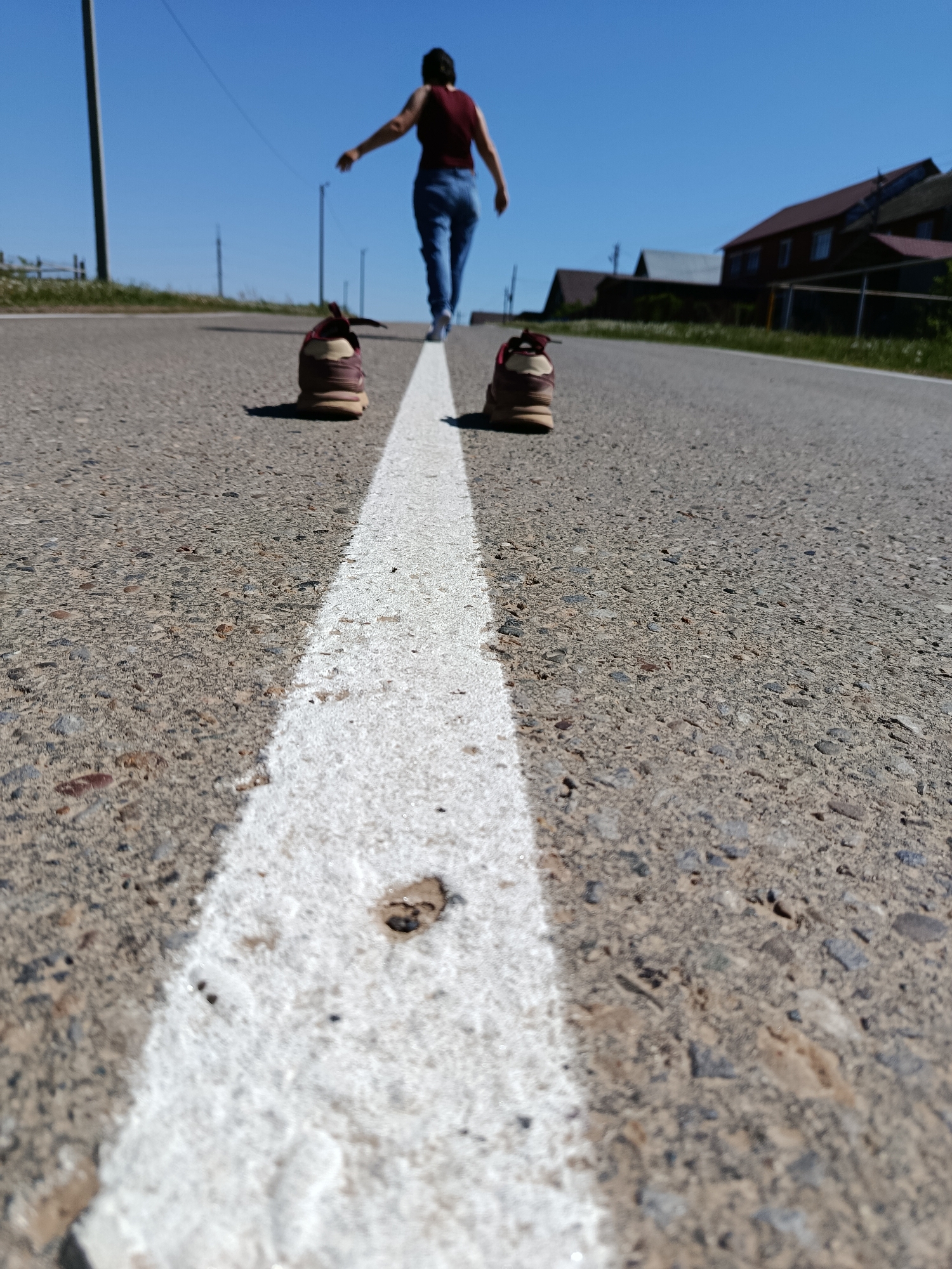A low-angle, medium shot depicts a pair of shoes on a road in Фоки, Russia. The shoes are positioned on either side of a thick, white painted line down the center of the asphalt road. The perspective of the shot creates a vanishing point in the distance. A woman, dressed in a red top and blue jeans, is walking barefoot away from the shoes down the center of the road toward the vanishing point. Houses are visible on the right side of the road and a telephone pole is on the left. The sky is a clear blue.