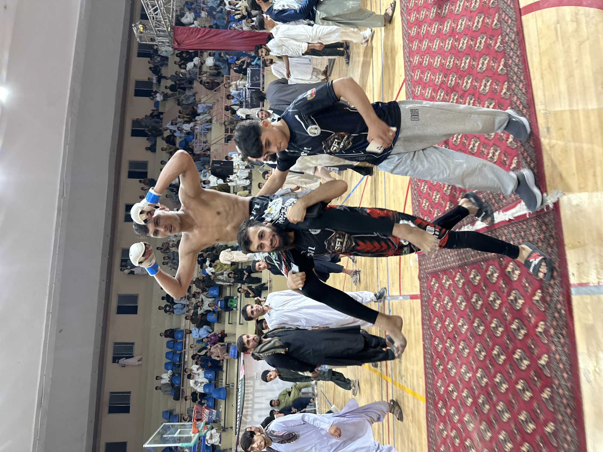 A group of three men are posing for a picture in an indoor stadium setting. The stadium appears to be filled with spectators seated in rows. The man in the foreground has a beard and is wearing a black and red shirt, holding the shirtless man on his shoulders. The shirtless man is wearing boxing gloves, and is smiling. The man behind him wears a black shirt with gray sweatpants, and he is holding a phone, possibly taking the photo. The stadium floor is partially covered by a traditional red rug with geometric patterns. Based on the presence of traditional clothing worn by the spectators, and the style of the rug, the location is most likely in Quetta City Tehsil, Pakistan.
