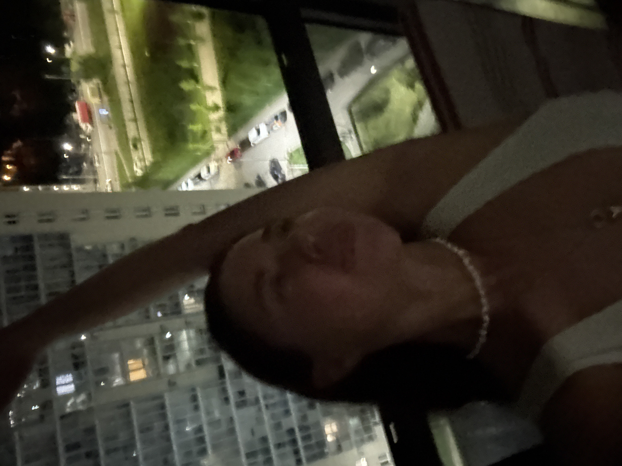 A person is positioned indoors, likely near a window, with a view of the city of Kazan, Russia, at night. The cityscape features illuminated buildings and streets with moving vehicles. The person is wearing a light-colored top and a necklace. The camera angle is looking down, possibly from an elevated vantage point.