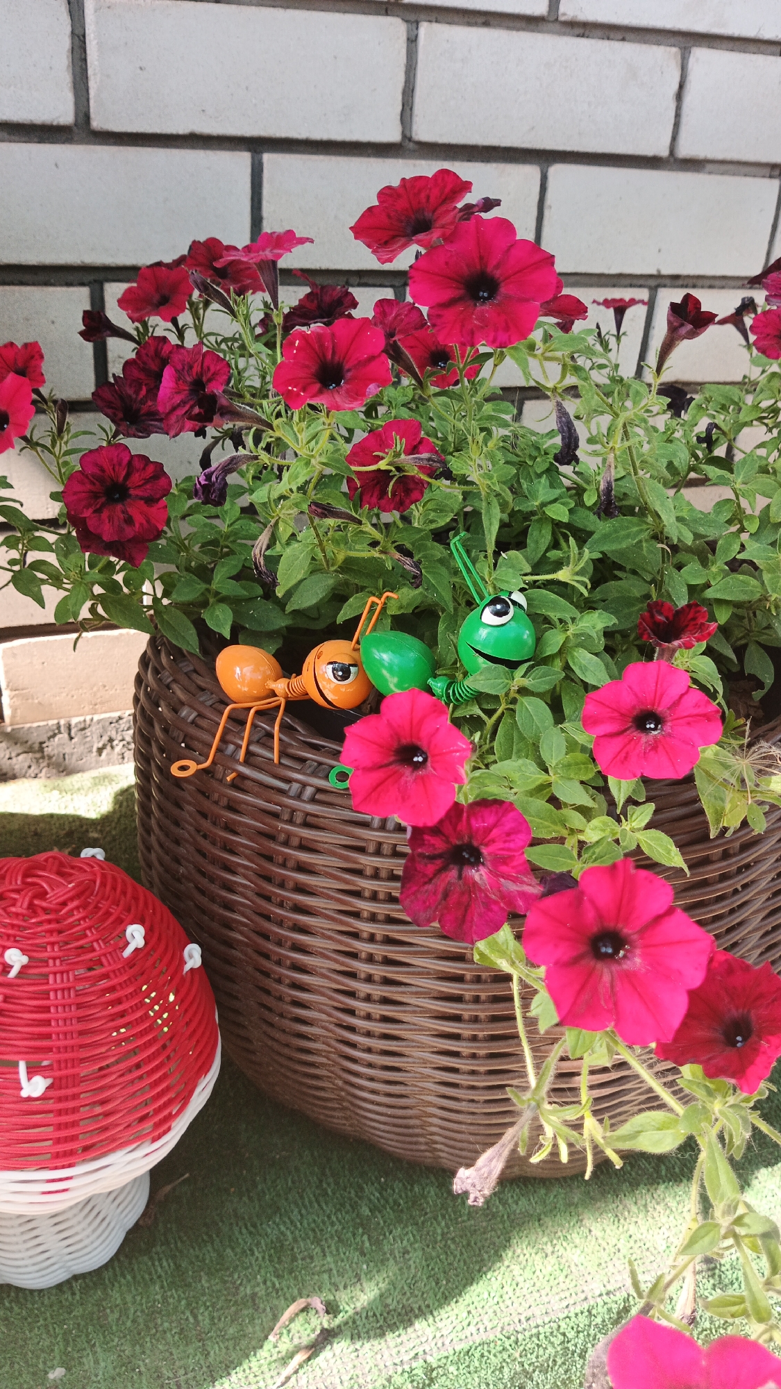 An outdoor, eye-level medium shot captures a decorative plant pot filled with vibrant pink and dark red petunias. The wicker pot rests on a green, artificial turf surface. To the left of the pot is a red and white decorative mushroom, also made of woven material. Two small, decorative insects, an orange ant and a green caterpillar, are placed on the edge of the pot, interacting with the flowers. The background features a light-colored brick wall. The scene is well-lit, suggesting it is daytime. No people are visible. The location is identified as Kryvyi Rih, Ukraine.