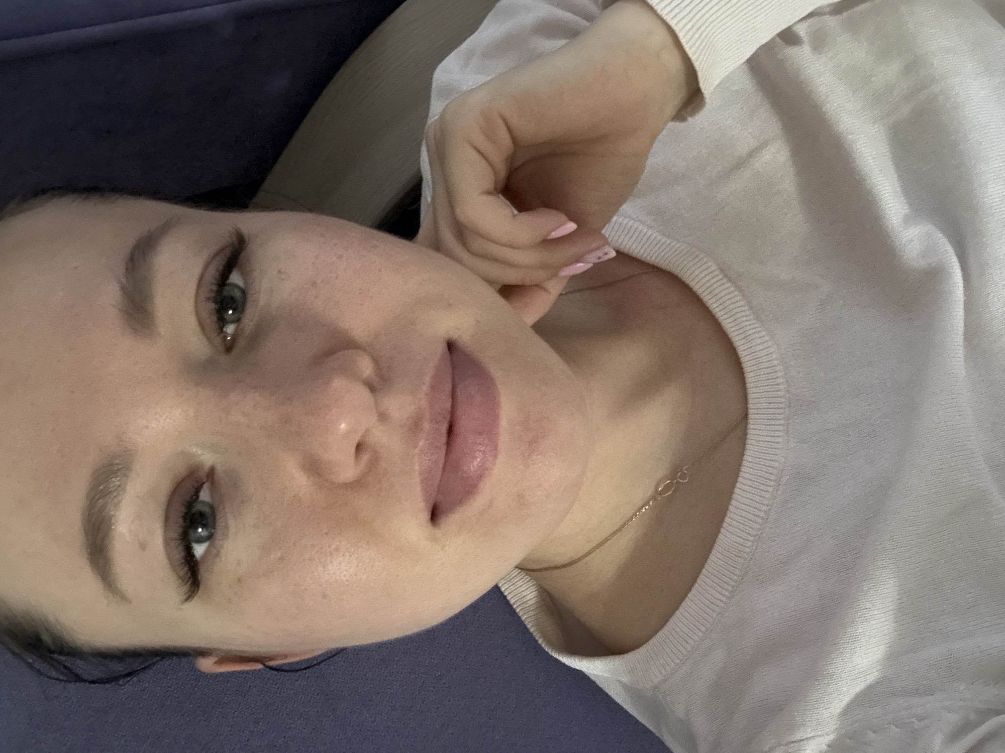 A close-up, tilted head-and-shoulders shot features a fair-skinned woman with light-colored eyes and long, dark eyelashes. She has full lips and freckles on her face. She is wearing a light-colored, round-necked shirt and a delicate gold necklace. Her left hand is raised, with her fingertips touching her cheek. She is sitting on a dark blue surface. The image was taken in Ферма, Russia.