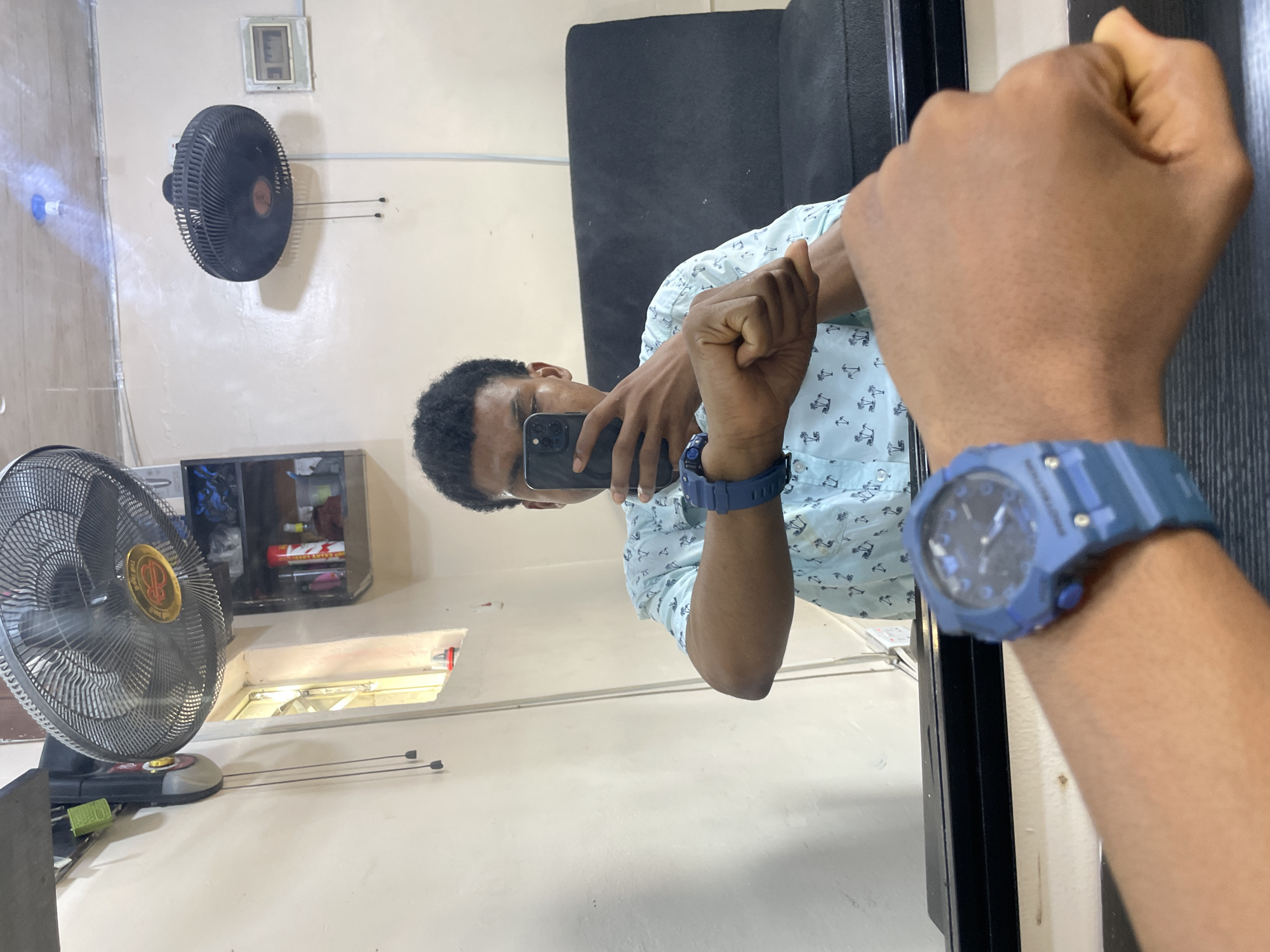 A medium-shot image depicts a man taking a mirror selfie indoors. The man is dark-skinned, wearing a light blue patterned shirt and a blue watch. He is holding a smartphone to capture his reflection.
The background features a wall-mounted fan and a standing fan, a cabinet containing various items. The location is likely in Ado, Nigeria, considering the prompt information.