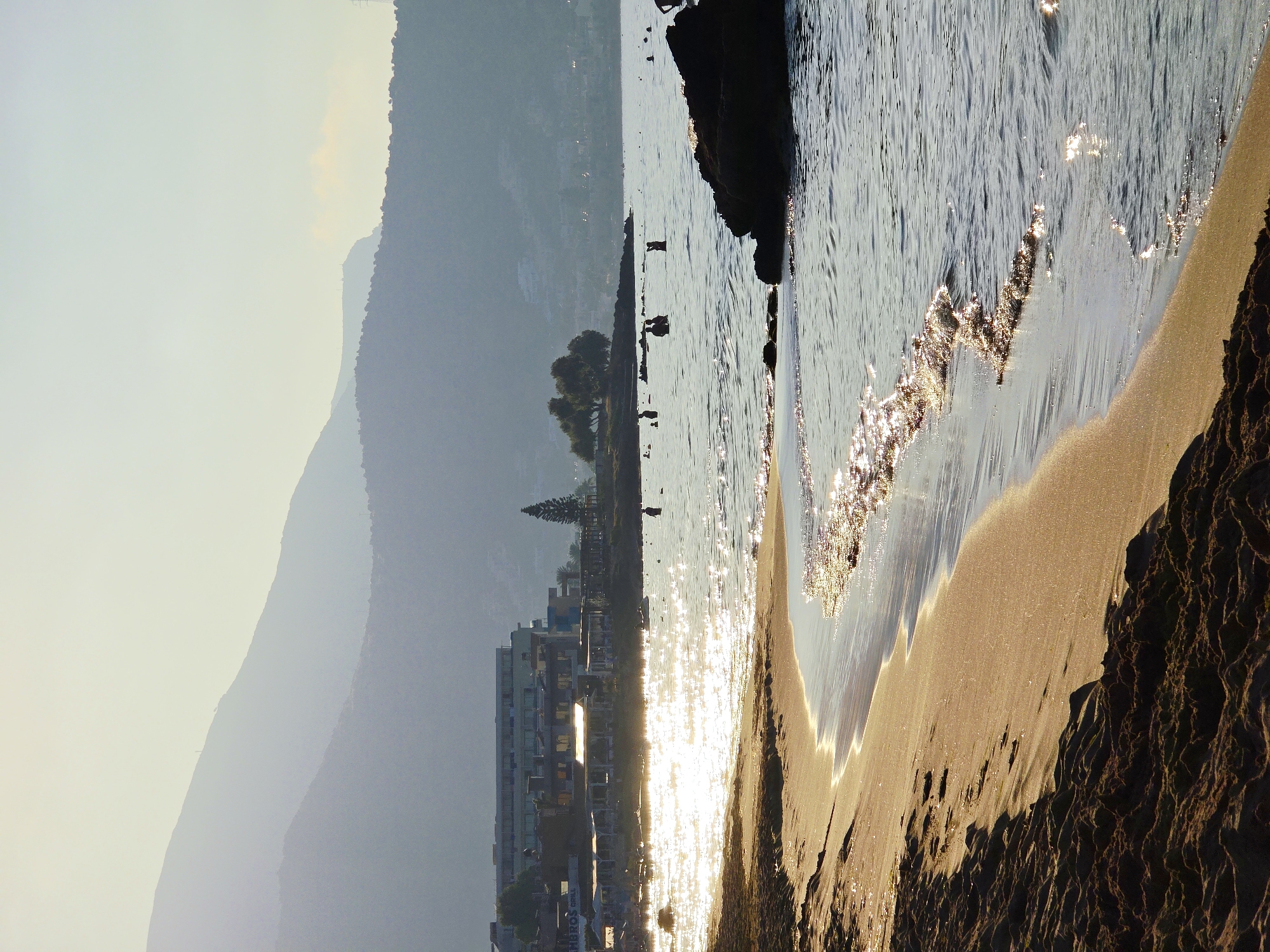 The image depicts a serene beach scene in Malia Municipal Unit, Greece, during the late afternoon or early morning. The foreground is dominated by a sandy beach with dark, textured sand in the lower left, transitioning to lighter, wet sand along the waterline. Gentle waves, glinting with sunlight, lap the shore. In the mid-ground, the sea is calm with scattered rocks and several figures swimming or wading. A prominent dark rock formation is visible on the right side. Further back, a row of buildings, appearing to be hotels or resorts, lines the coast, with some structures featuring awnings or outdoor seating areas. A lone pine tree stands near the buildings, and a few more trees are scattered along the coastline. The background features hazy, layered mountains that fade into the pale, clear sky, with a subtle hint of a settlement on the slopes of the furthest mountain. The overall atmosphere is peaceful and idyllic, bathed in the soft, warm light of the sun.