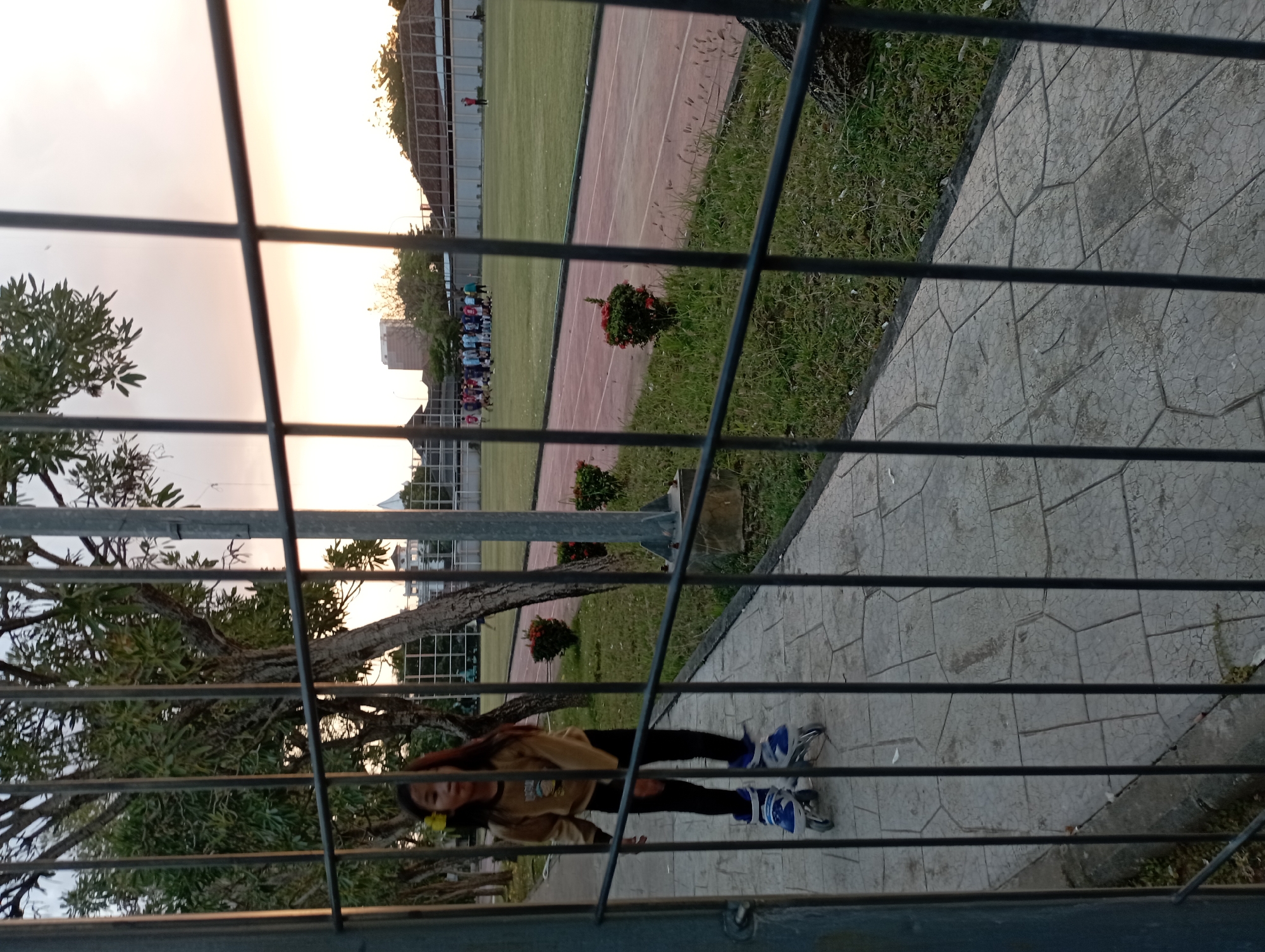 A young girl with long, dark hair and a yellow flower in her hair is rollerblading on a paved walkway. She is wearing a long-sleeved brown shirt and black pants. The walkway is bordered by a grassy area with bushes and trees. In the background, there is a sports field with a track and a group of people, likely playing a sport. Several buildings are visible in the distance, and the sky is a pale orange, suggesting it is either sunrise or sunset. The scene is viewed through a metal fence. This is likely located in Surakarta, Indonesia, given the vegetation and architectural style.