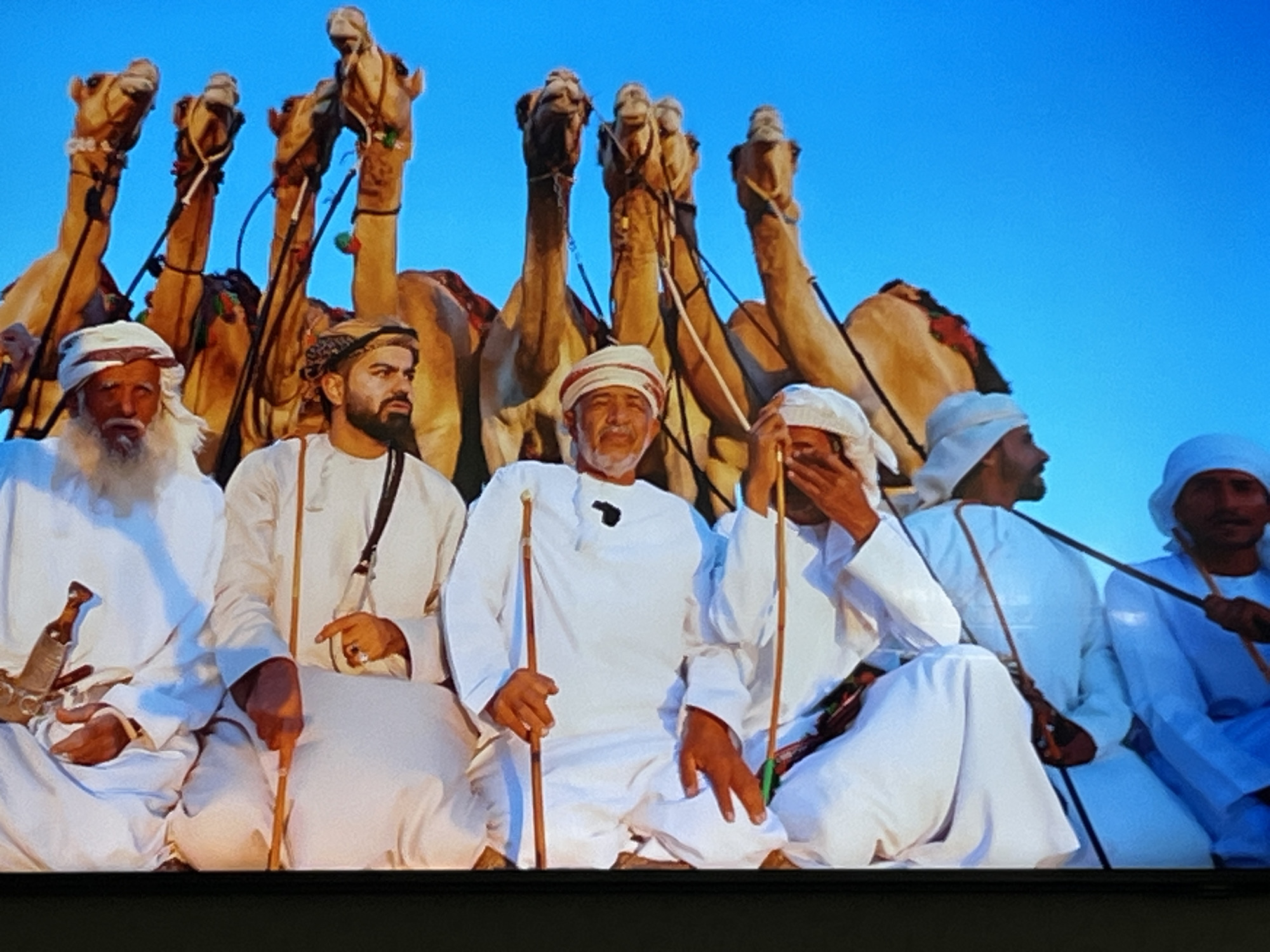 The image shows a group of men in traditional Omani clothing, seated in front of several camels. The men are wearing white thobes and headscarves. Some of the men are holding walking sticks. The camels are standing behind the men, and their heads are visible above the men's shoulders. The sky is a clear blue. There is no discernible indication of the city of Kharkiv, Ukraine.