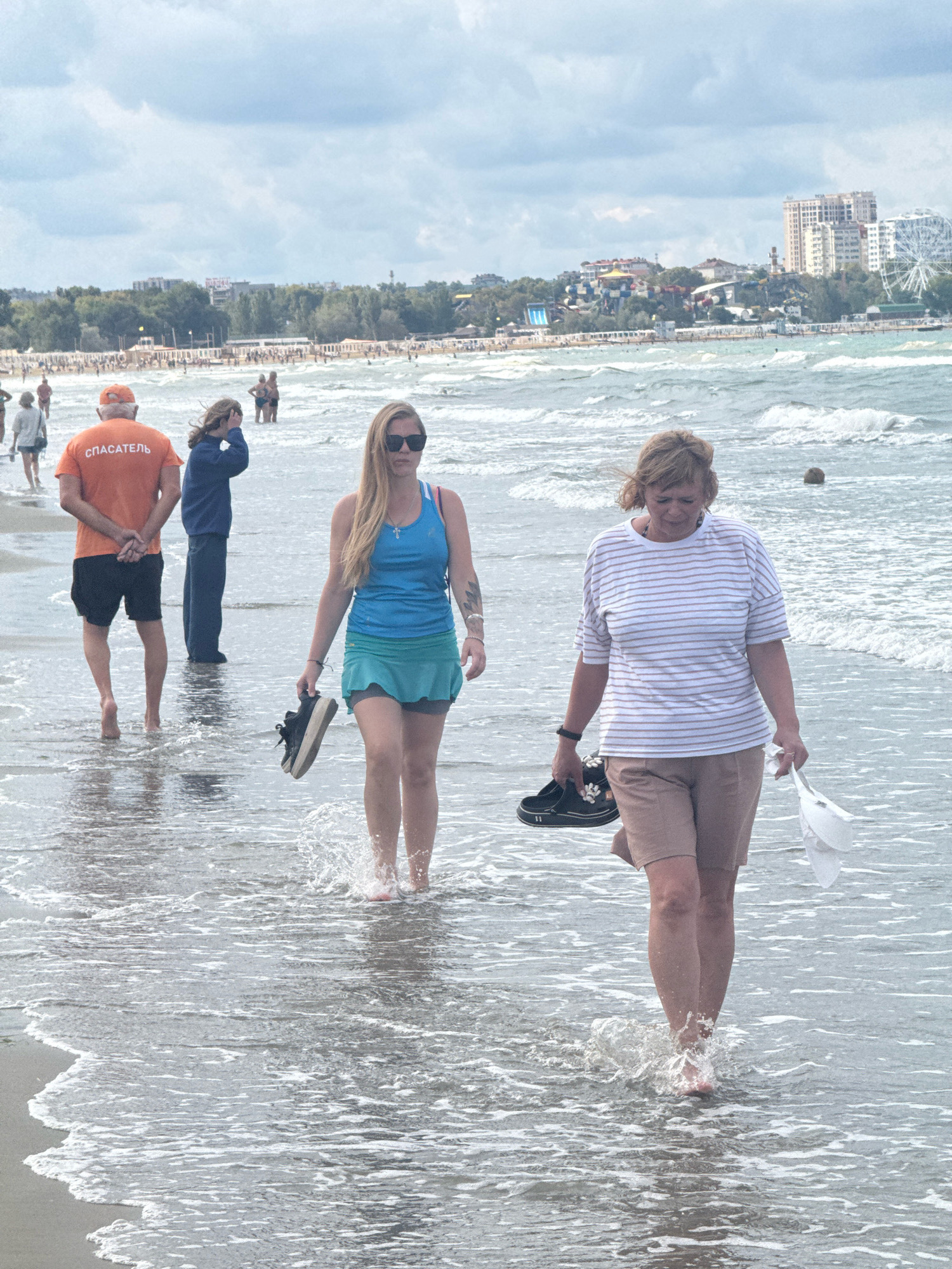 Anapa, Russia, is pictured on a cloudy day at the beach with the Black Sea in the foreground. Four individuals are visible walking on the wet sand near the water's edge.
A man in an orange shirt with the word "СПАСАТЕЛЬ" (Lifeguard) in white Cyrillic lettering, black shorts, and an orange cap is walking away from the camera. His hands are clasped behind his back. To his left and slightly behind him, a person in a dark blue hooded jacket and dark pants walks with their left hand to their head.
In the foreground, a woman with blonde hair and sunglasses is walking towards the camera, holding a pair of black sneakers in her right hand. She is wearing a bright blue tank top and a short teal skirt. To her right and closer to the camera, a woman with blonde, short hair is walking towards the camera, carrying a white bag or hat in her right hand and a pair of dark sneakers in her left. She is wearing a white horizontally striped t-shirt and light brown shorts.
The sea has moderate waves, and there are people and structures visible on the distant shoreline, including buildings, a Ferris wheel, and what appear to be water slides. The sky is overcast with scattered clouds.