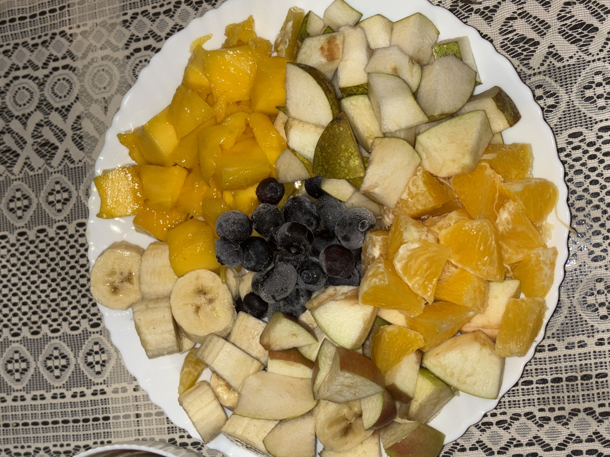 A white plate of sliced fruit is centrally located on a patterned, neutral-toned tablecloth. The plate contains chunks of mango, pear, oranges, frozen blueberries, and sliced banana. The scene appears to be indoors. The exact location in Vnukovo, Russia cannot be determined from the image.