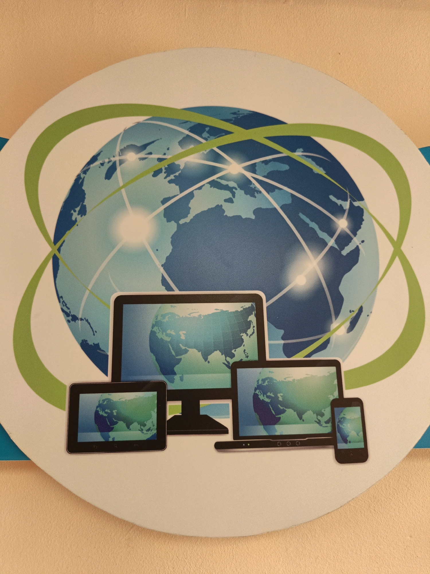 A close-up shot shows a circular sign with an illustration. The sign features a blue globe with light blue continents, surrounded by green orbit lines. A series of digital devices are centrally located on the lower half of the sign, including a desktop computer, a tablet, a laptop, and a smartphone. Each device displays a map of the Earth. The sign appears to be mounted on a plain, light tan wall. There is no visible evidence to confirm that this is from Парэчча, Belarus.