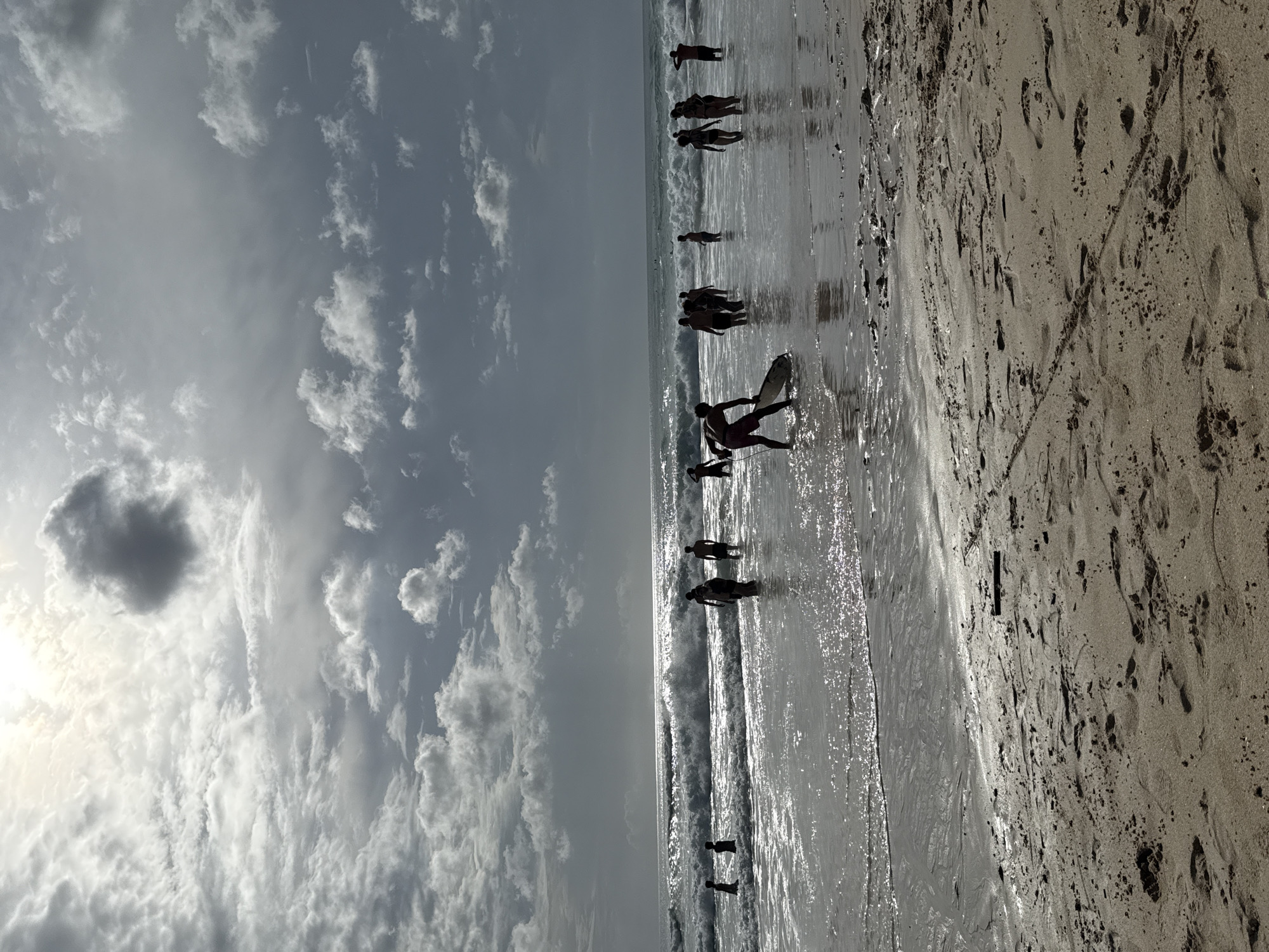 The image depicts a beach scene, likely in Kuta Selatan, Indonesia, given the context. The view is perpendicular to the shoreline.
Sky: The sky occupies roughly half the image and is filled with a mix of dense, grey-white clouds. Some clouds are more diffuse, while others have a darker, denser appearance. The sun appears to be obscured by the clouds, creating a bright spot in the sky.
Ocean and Shoreline: The ocean meets the sandy beach. Several individuals, appearing as silhouettes, are seen wading in the shallow water. One person appears to be carrying a surfboard. The ocean surface is somewhat reflective, with visible ripples and small waves.
Beach: The sandy beach occupies the other half of the image and shows numerous footprints and scattered debris. The sand color varies from light beige to darker shades, possibly due to moisture content.