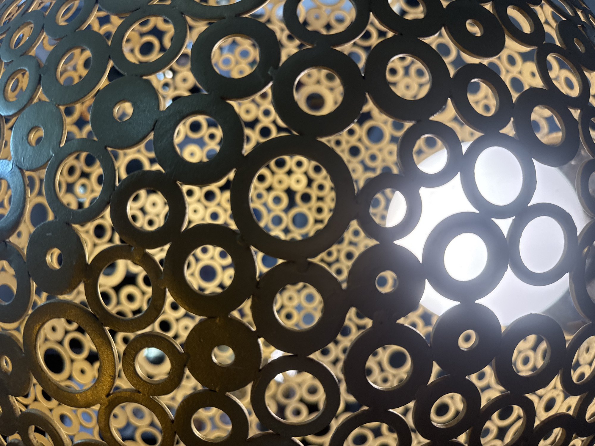 A close-up shot reveals a spherical lighting fixture or decorative object. The object is constructed of interconnected metallic rings of varying sizes. In the foreground, larger rings with a gold or brass-like finish are prominently displayed. Behind these, a dense pattern of smaller rings is visible, creating a textured backdrop. The light source within the object is bright and diffuse, emanating through the spaces created by the rings. There is nothing that suggests that this image was taken in Nha Trang Ward, Vietnam.