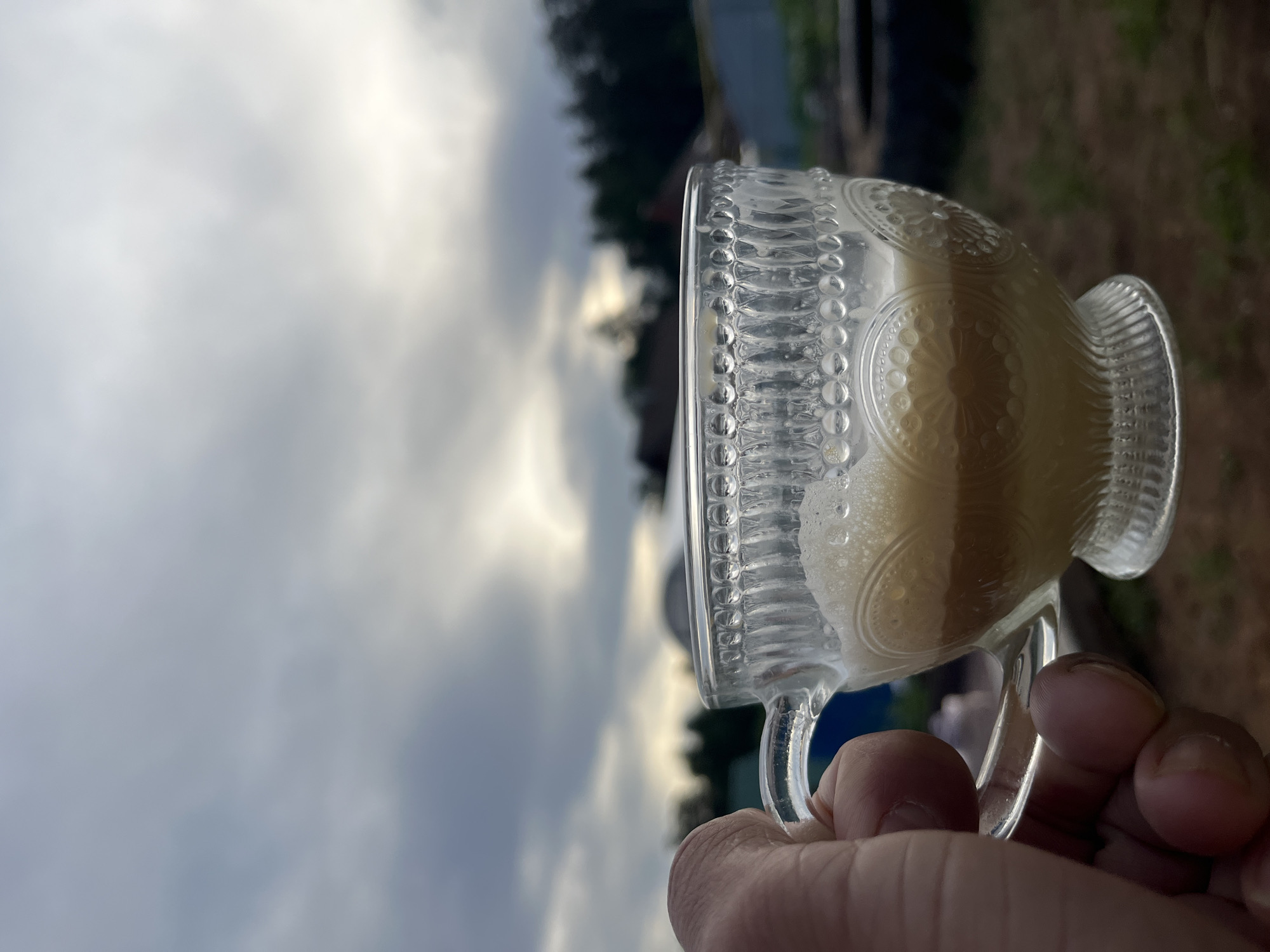 A hand holds a glass cup filled with a light tan beverage, likely outside. The cup has a decorative, vintage design. Overcast skies and trees are in the background. The location cannot be confirmed as Chaikovsky, Russia.