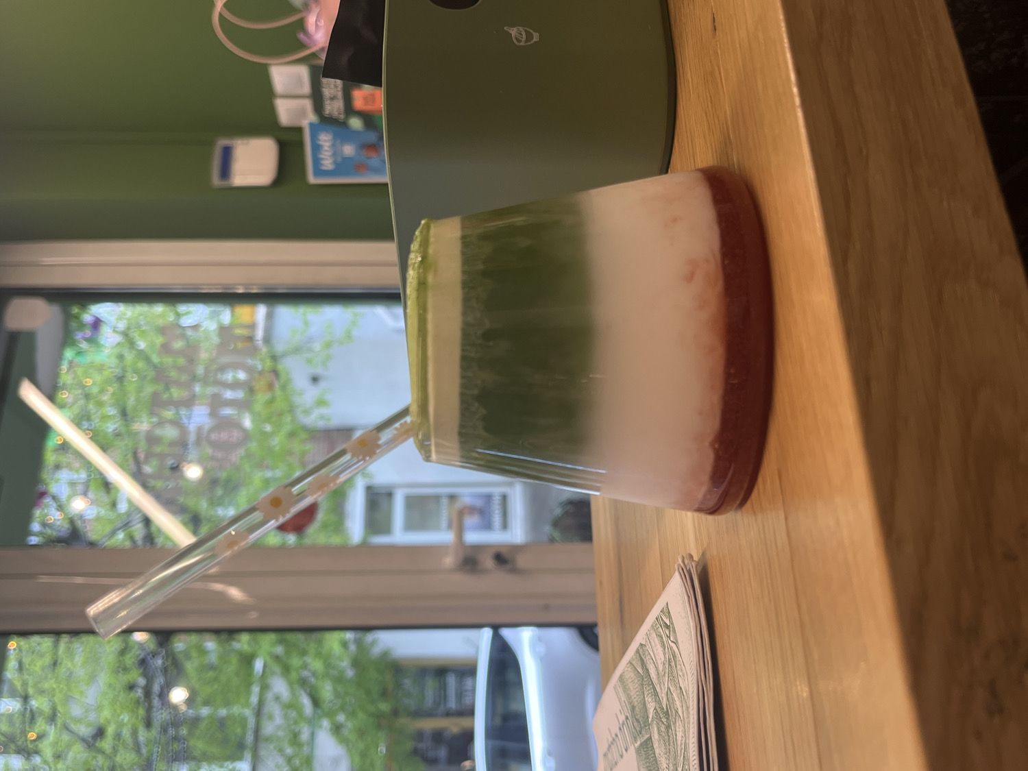 The image, when viewed upright, features a clear glass holding a layered beverage, placed on a light-colored wooden table. The drink displays three distinct horizontal layers: a vibrant green at the top, a milky white in the middle, and a deep reddish-brown at the bottom, possibly indicating a matcha, milk, and fruit puree combination. A transparent straw, decorated with delicate white daisy patterns, is inserted into the glass.
On the wooden table to the left of the drink, a stack of papers or menus, partially covered, shows subtle green illustrations. The table surface is clean and smooth, highlighting its natural wood grain.
The background reveals a window looking out onto a blurred outdoor scene with lush green trees and foliage, suggesting a pleasant daytime setting. The natural light filtering through indicates it is not deep night. Above and to the right of the window, an interior green wall is visible. Mounted on this wall are a white rectangular object, possibly an intercom or dispenser, and a distinctive blue sign. The word "Wolt" is clearly visible on this blue sign, indicating the presence of a food delivery service affiliation.
The scene conveys a calm atmosphere in a cafe or similar establishment, consistent with a modern urban setting like Sofia, Bulgaria. No people are visible in the frame, keeping the focus on the beverage and its immediate surroundings.