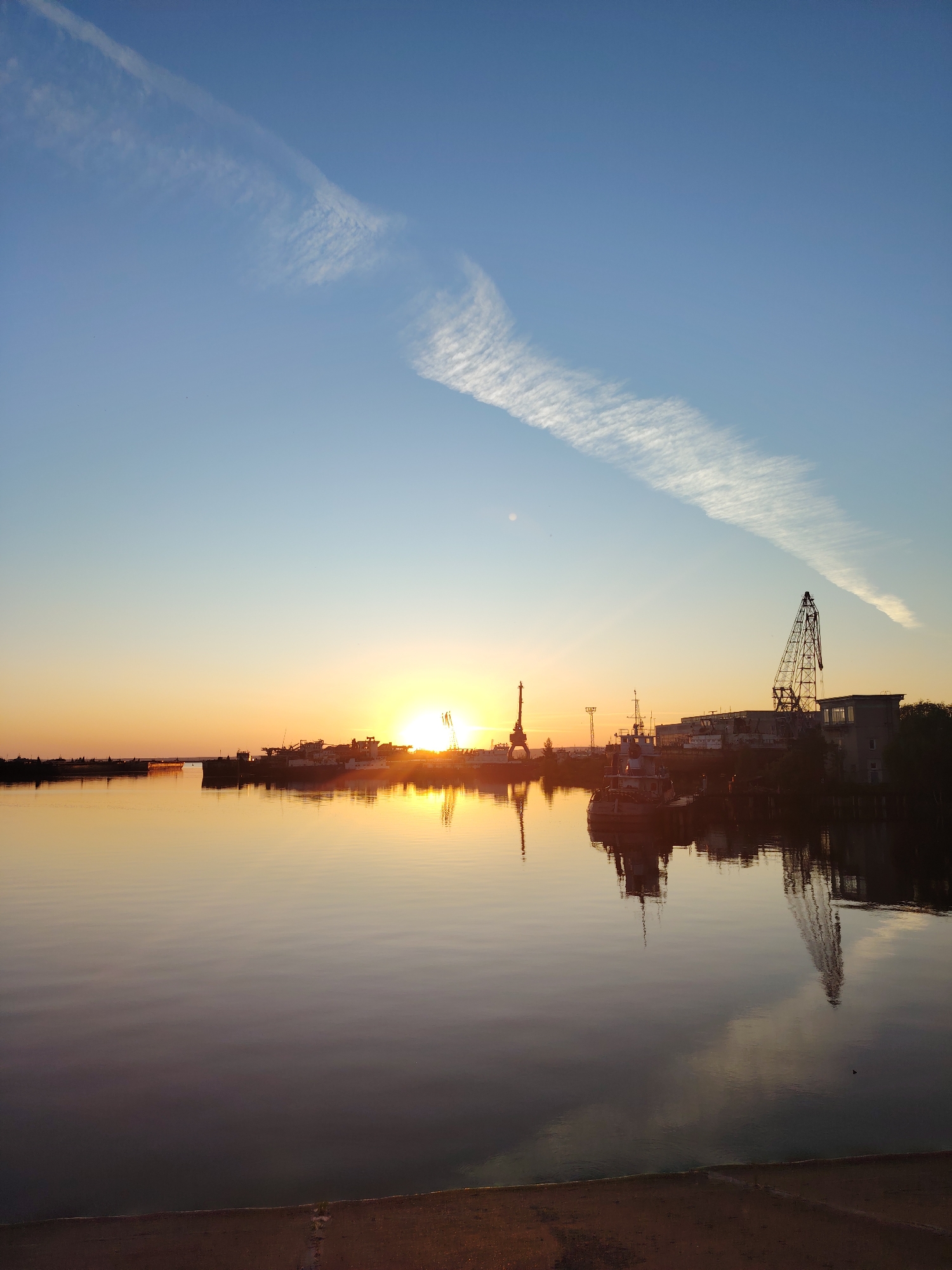 The image captures a tranquil scene at sunrise in the city of Chaikovsky, Russia, along the Kama River. The sky is a gradient of soft blue fading into warm orange and yellow hues near the horizon, with a wispy cirrus cloud streak traversing the upper left portion. Silhouetted against the colorful sky are several industrial structures and vessels docked along the riverbank.
Prominent among these are industrial cranes, including a large, skeletal tower crane on the right side of the image and another to the left, closer to the sun. Several ships and boats are moored, their forms indistinct in the low light. A larger building with windows is visible behind the cranes on the right.
The calm surface of the Kama River acts as a mirror, reflecting the sunrise colors and the silhouettes of the ships and cranes, creating a symmetrical and peaceful composition. The foreground shows a strip of sandy embankment, contrasting with the smooth water. There are no people visible in the frame, and the overall atmosphere is one of quietude and the start of a new day.
