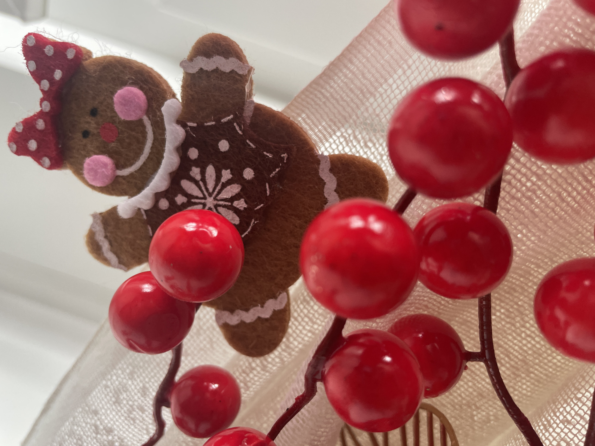 The image shows a close-up of a Christmas decoration. A gingerbread woman made of felt is visible on the left side. It has a red and white polka dot bow, pink cheeks, and white trim around the neck and waist. To the right, there are several bright red, glossy berries clustered together on brown stems. A cream-colored, textured fabric serves as a backdrop. There is no information in the picture to suggest it was taken in Novi Sad, Serbia.