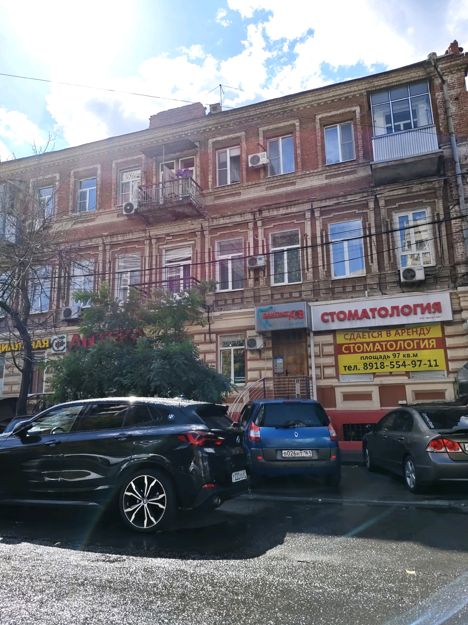 The image depicts the facade of a brick building in Rostov-on-Don, Russia. The building has multiple stories with several windows, some of which are air-conditioned units. There are balconies on the upper floors. Signs are visible on the building, including "СТОМАТОЛОГИЯ" (Dentistry) and "СДАЕТСЯ В АРЕНДУ СТОМАТОЛОГИЯ" (Dentistry for rent). Several cars are parked on the street in front of the building. A large green tree is in front of the building, partially obscuring some of the lower-level signs. The sky is visible with clouds and sunlight, indicating it is daytime.
