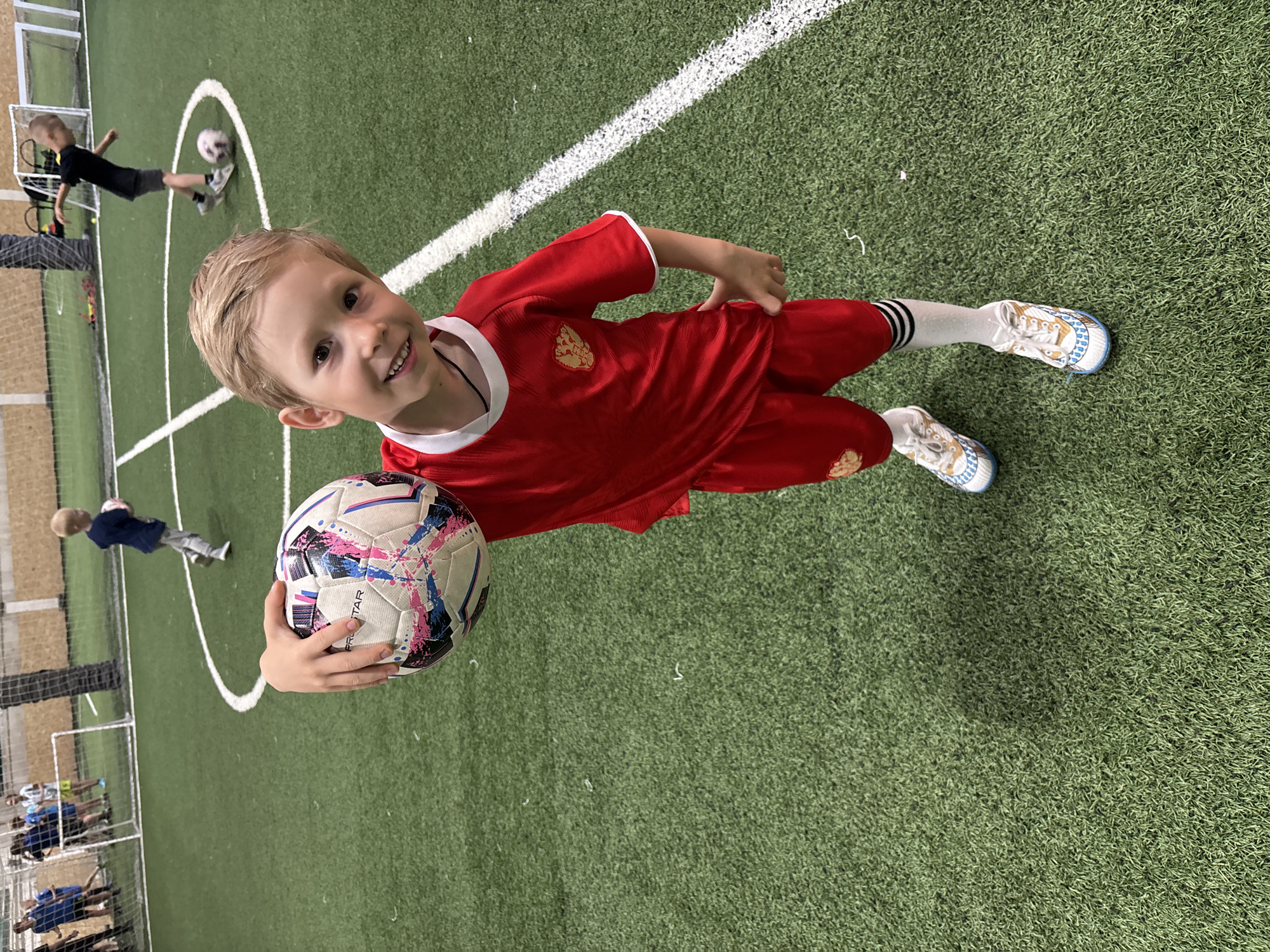 A young, fair-skinned boy with blonde hair smiles and holds a soccer ball in his left hand. He is wearing a red soccer uniform with white trim. The scene appears to be indoors on a soccer field with artificial green turf, indicated by the grainy texture. A white line is visible in the background. Other individuals are present in the background, some engaged in soccer-related activities. The image suggests the location is a soccer facility, potentially located in Chaikovsky, Russia.