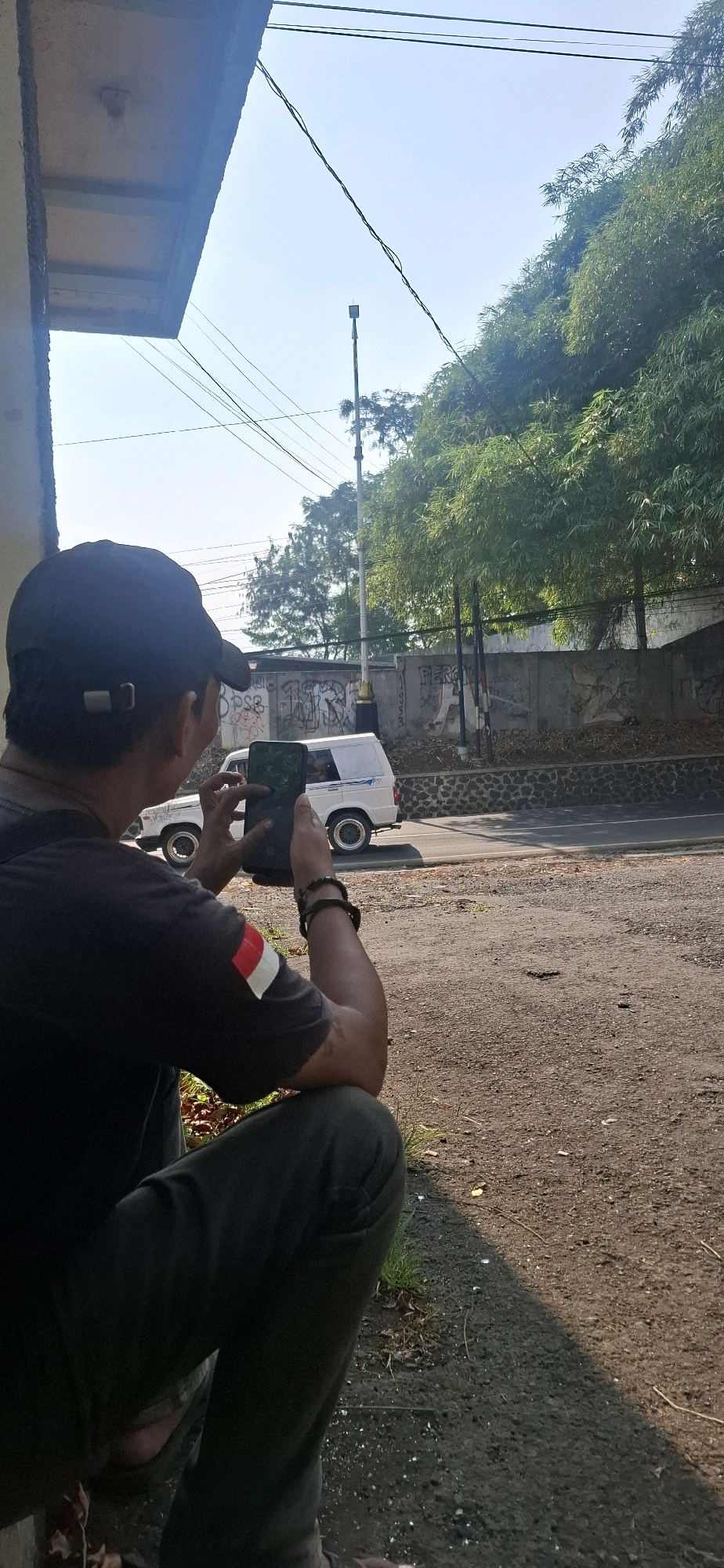 An over-the-shoulder, vertical, outdoor, eye-level medium shot captures a person wearing a dark t-shirt with a red and white stripe on the left sleeve and a dark baseball cap. The person is crouched down, holding a smartphone in both hands, appearing to record or photograph something off-camera to the right. The person's left arm is bent, with a bracelet on the wrist. The background features a gravelly ground, a road with a white van driving on it, a stone wall with graffiti, and lush green trees, possibly bamboo, with a clear blue sky. A lamppost is visible in the distance. The scene is consistent with an outdoor urban or semi-urban environment in Cilaku, Indonesia, likely a roadside area given the presence of the road and wall. The lighting suggests it is daytime, possibly morning or afternoon, with sunlight casting shadows on the ground.