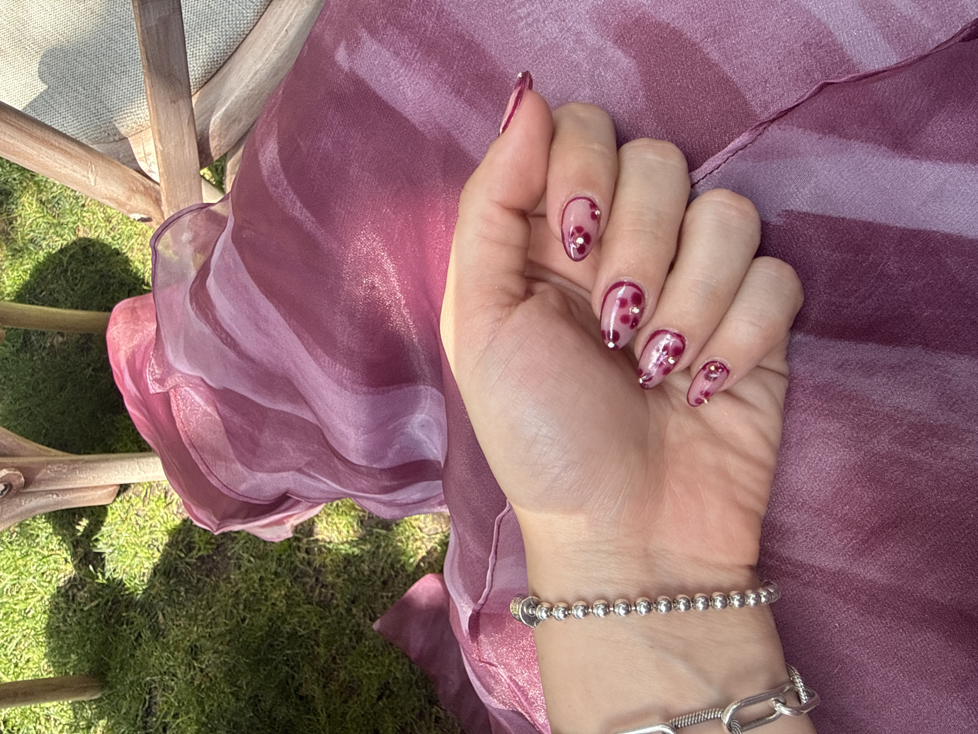 A hand rests on a rose-colored fabric. The hand is adorned with two silver bracelets. The nails are painted in a pale pink with burgundy accents and small silver embellishments. The fabric is draped over a wooden chair. In the background is green grass, partially in shadow. The location is likely outdoors. There is no definitive evidence in the image to confirm the location is Santiago de Querétaro, Mexico.