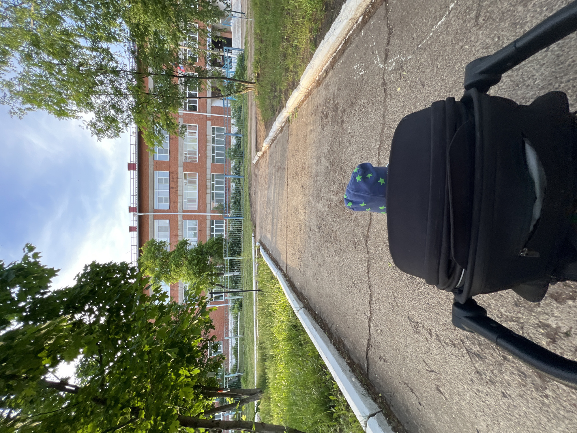 The image is a first-person view from behind a black stroller, with a child in blue star-patterned clothing visible inside. The stroller is on a paved pathway with a white curb. To the left of the path is a grassy area with trees and a metal fence. Beyond the fence is a multi-story brick building with white window frames and a red roof. The sky is partially visible with some clouds. The location is Noviy, Russia.