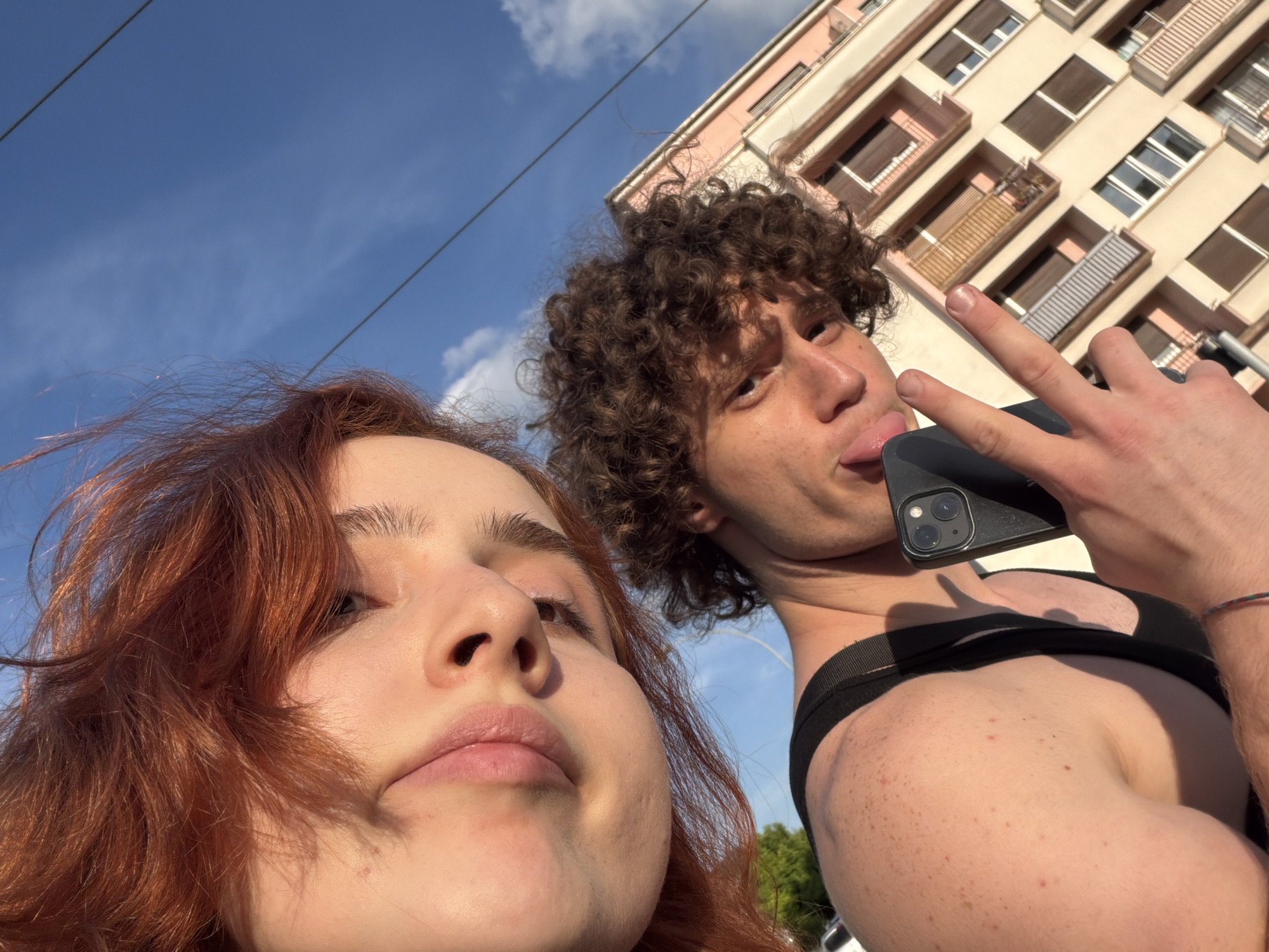 This low-angle outdoor selfie captures two individuals, a woman and a man, under a clear blue sky with sparse white clouds. In the foreground, partially cropped, is the profile of a woman with reddish-brown hair, her face illuminated by bright, warm sunlight. She appears relaxed, looking slightly upwards.
Behind her and to the right, a man with curly brown hair gazes directly at the camera with a playful expression, his tongue extended slightly. He forms a peace sign with his left hand, which also holds a black smartphone, partially obscuring his lower face. He is wearing a black tank top or undershirt.
The background reveals a multi-story urban building with a light-colored facade, featuring numerous windows, some with closed blinds, and balconies. The architecture suggests a European city setting, consistent with the context of Strasbourg, France. Overhead wires are faintly visible crossing the sky. The strong sunlight and shadows indicate a bright daytime scene, likely early morning or late afternoon. There is no visible text in the image.