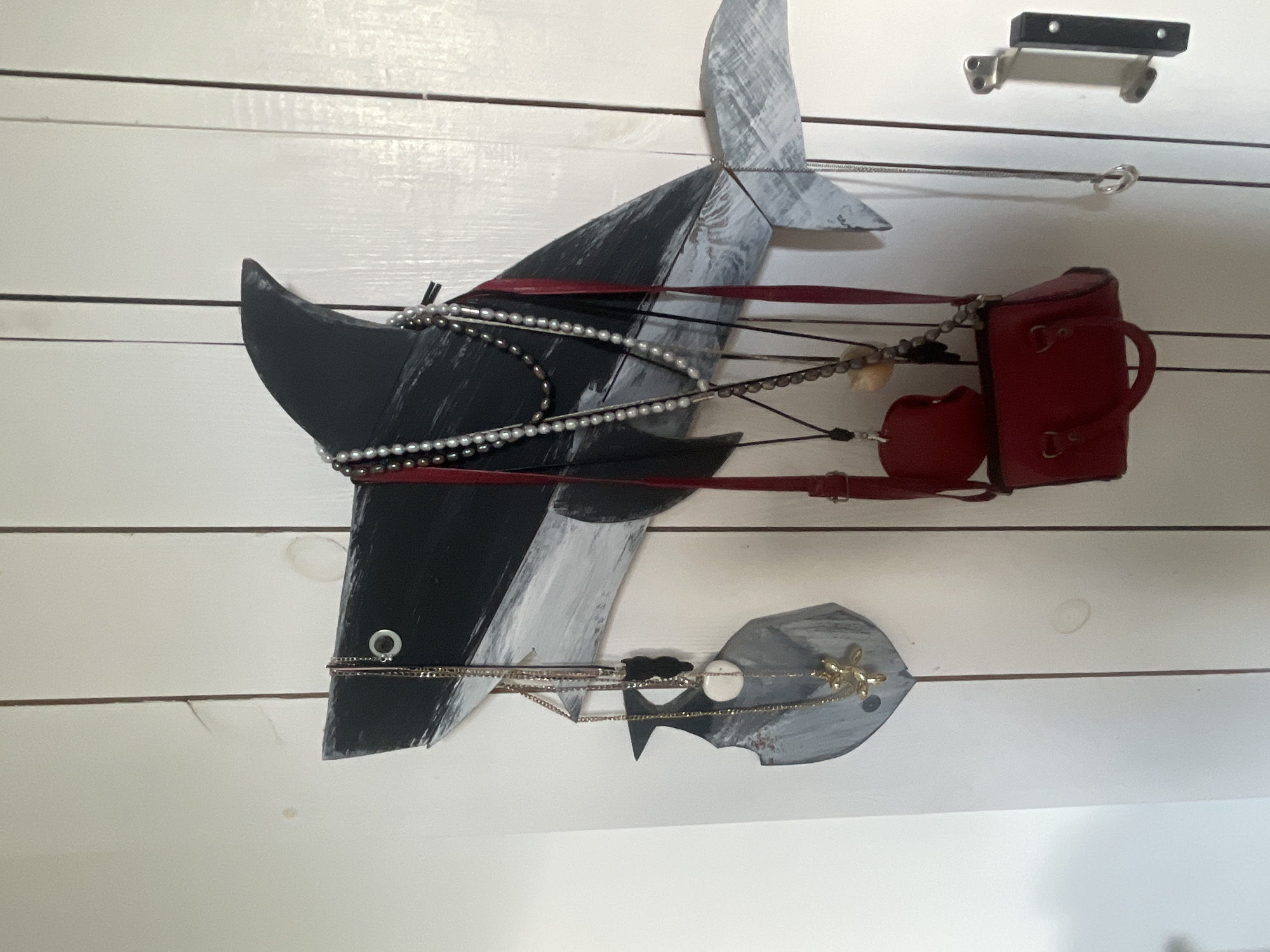 A wooden sculpture of a shark is mounted on a white wooden plank wall. The shark is painted in black and gray, with its mouth open, revealing a red interior. Dangling from the shark are strands of pearls and chains, as well as a small red handbag, a round beige object, and a heart-shaped red charm. The shark's tail is a separate piece, painted in gray and attached to the body by thin wires. Below the shark is another wooden piece, also painted gray, resembling a fin or a smaller fish, adorned with a golden, balloon-dog-shaped charm. The overall presentation suggests a whimsical decorative item, perhaps hung indoors. The lighting is even, indicating artificial light or an overcast day. There is no visible text in the image. The context provided is "Мухавецкі сельскі Савет, Belarus," which might indicate the general region where this item is located or was found.