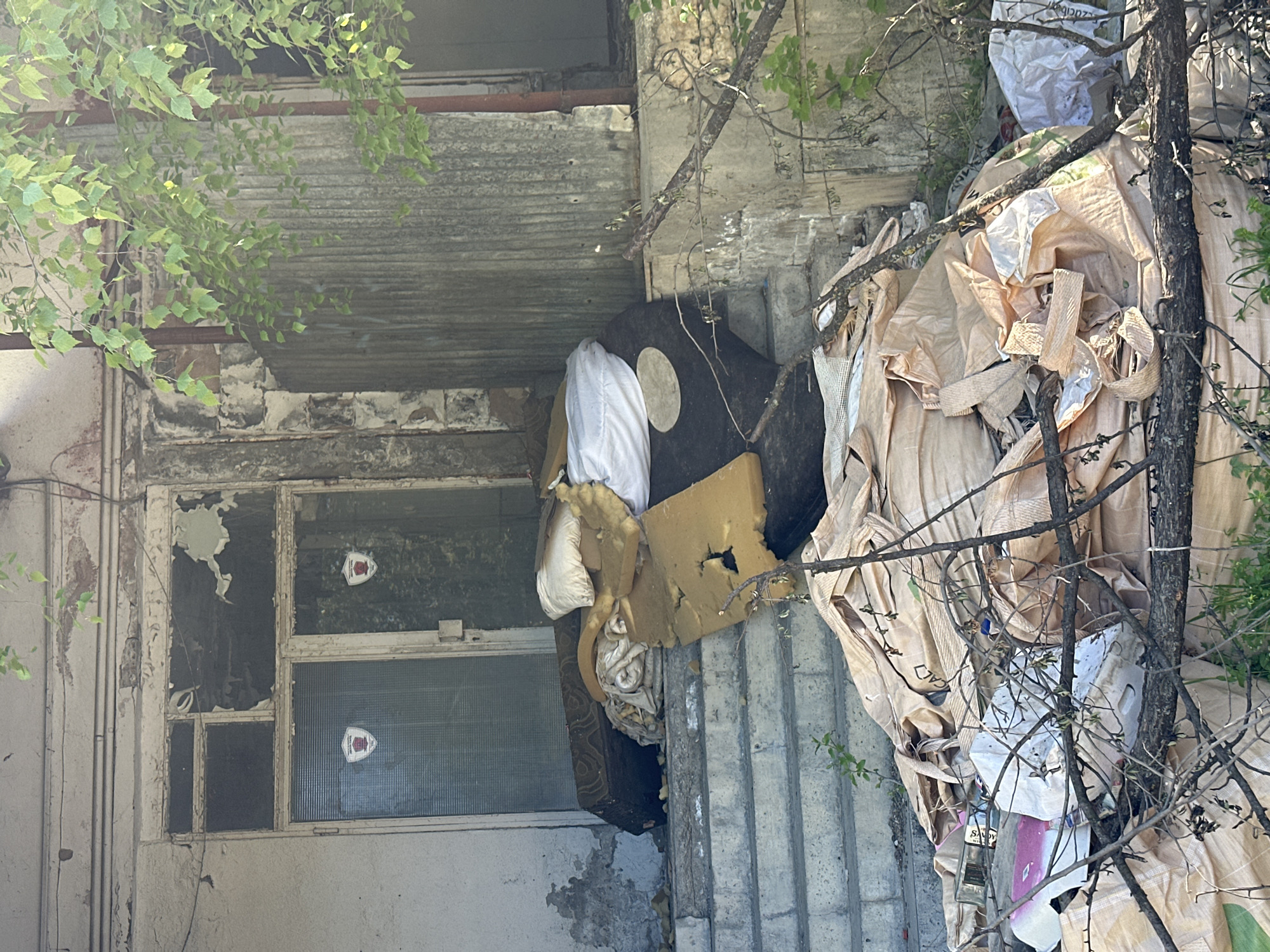 The image captures a scene of significant urban decay and accumulated refuse next to a dilapidated building in Lovech, Bulgaria, viewed in bright daylight. The setting is characterized by neglect and overgrowth.
On the left, a weathered building facade is visible, with peeling light-colored paint revealing darker layers and concrete beneath. A window frame, likely from an old commercial or residential structure, contains a broken pane opening to a dark interior. Adjacent to it, another window with ribbed glass has two small, white and red shield-shaped stickers.
The right side of the frame is dominated by a large, disorderly pile of discarded items. This refuse includes numerous large, industrial-style bags in shades of beige and light brown, some appearing torn. Faint, fragmented text, such as "CACI," is barely discernible on some bags. Interspersed with these bags are various pieces of what appear to be old furniture or upholstery, including sections of yellow foam padding, a dark, patterned cushion with a circular light patch, and bundles of white fabric. A clear glass bottle, with the text "SANDY" partially visible on its label, is also present among the debris. Tree trunks and branches, some with green leaves, emerge from and intertwine with the pile, indicating that the waste has accumulated over a long period.
No people are visible, and there is no discernible activity taking place, reinforcing the sense of abandonment. The clear light suggests a sunny day. The overall impression is one of environmental neglect and urban blight.
