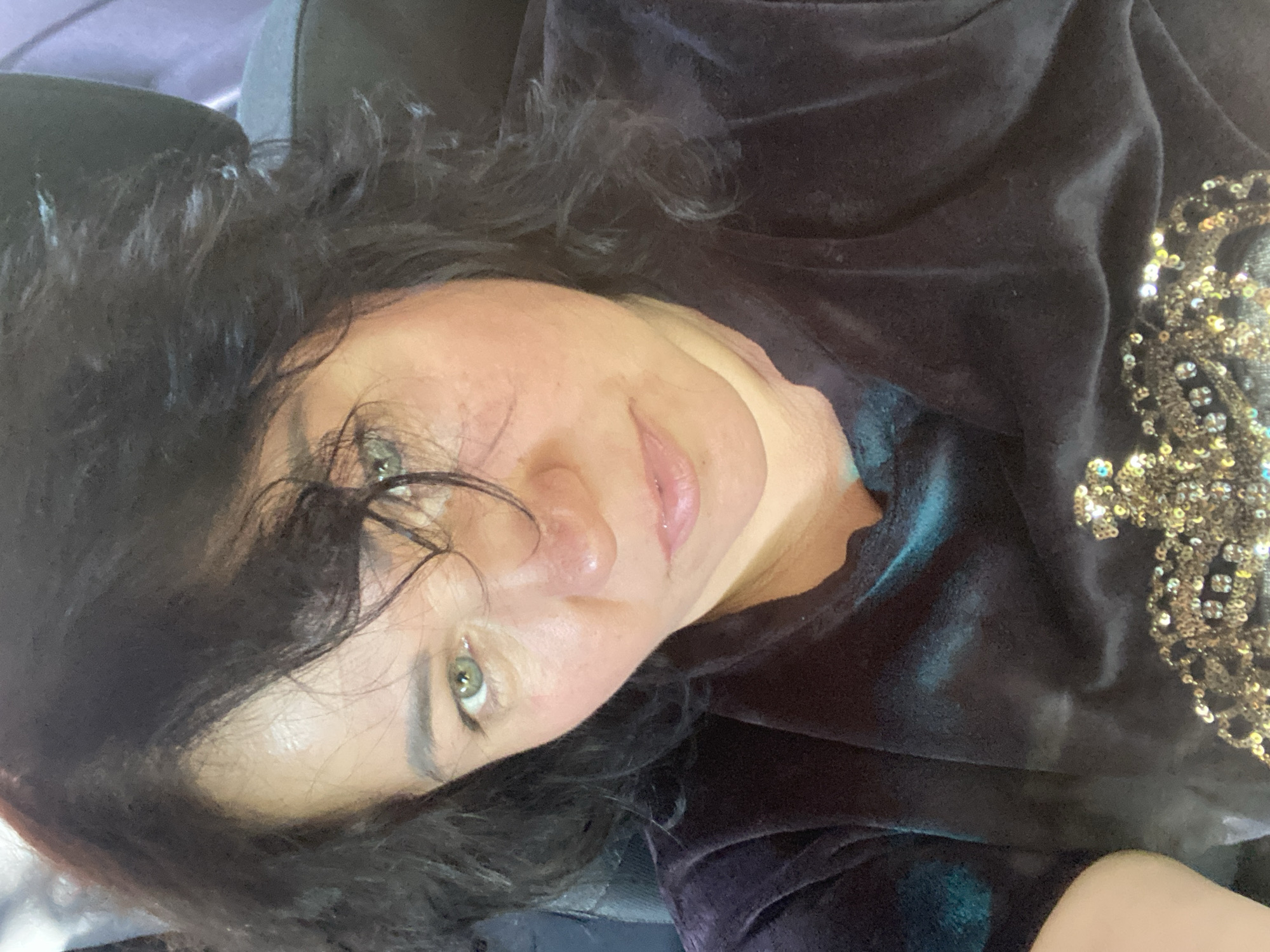 The image is a selfie of a woman with dark, wavy hair and green eyes. She is wearing a dark velvet top with an ornate, glittering design on the chest. Her skin appears flushed, and there is some redness around her nose. The background is out of focus but appears to be an interior setting, possibly a car or public transportation with a purple headrest visible. The lighting suggests it is daytime. The image does not contain any information that would allow for the determination of its location in the city of Dnipro, Ukraine.