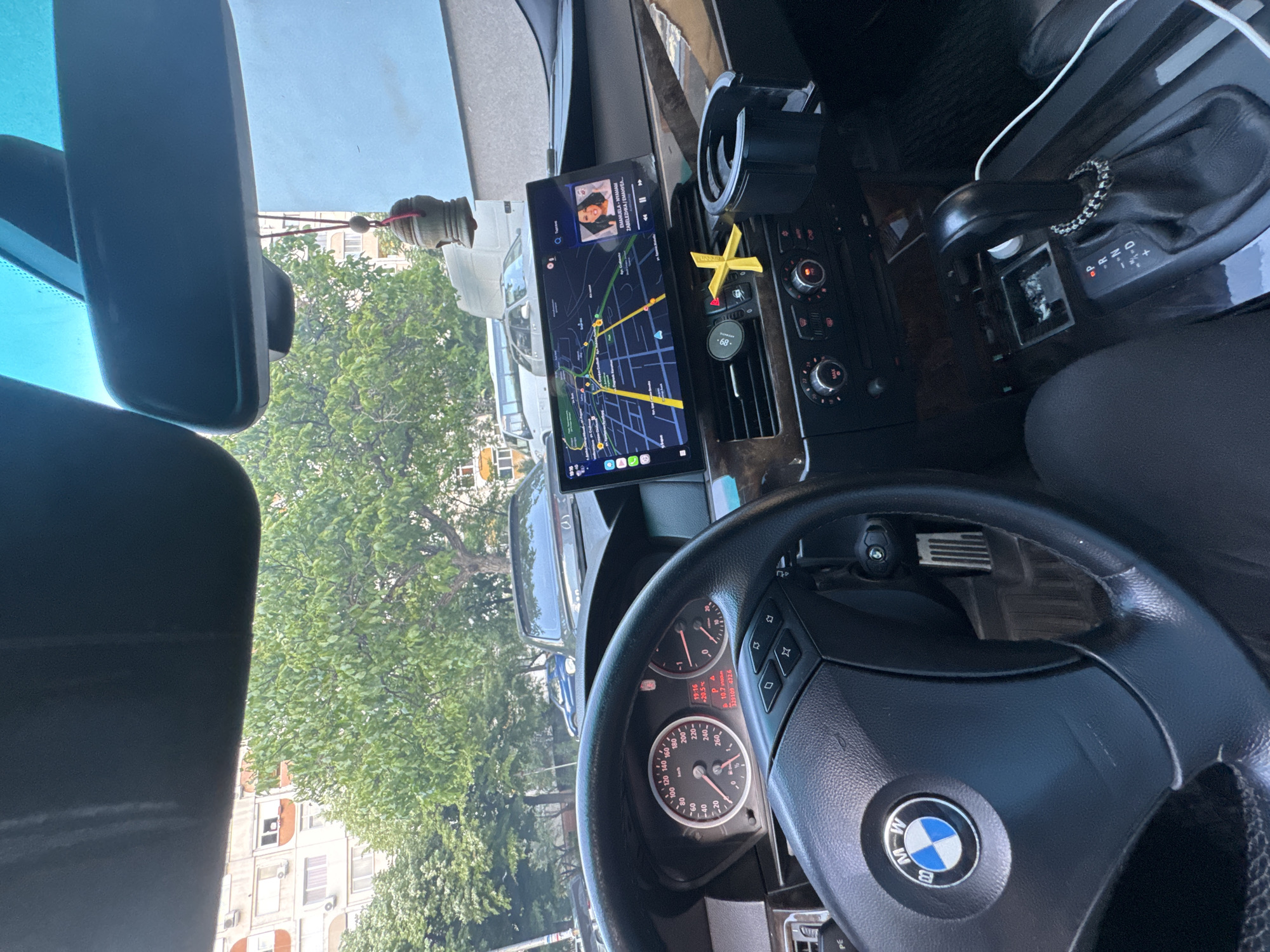 The image depicts the interior of a BMW vehicle, specifically the driver's area. The focus is on the dashboard, steering wheel, and central console. The infotainment screen displays a navigation map of what appears to be the city of Stara Zagora, Bulgaria. The steering wheel features the BMW logo. The dashboard includes controls for climate settings, as well as a cup holder. The car is parked facing a street with trees and buildings in the background. The gear shift is visible on the console. The speedometer shows 0 km/h. A car freshener is clipped onto the air vent. A decorative beaded ring is on the gear shift. An item hangs from the rearview mirror. The time is 19:16 and the temperature is 20.5 degrees Celsius.