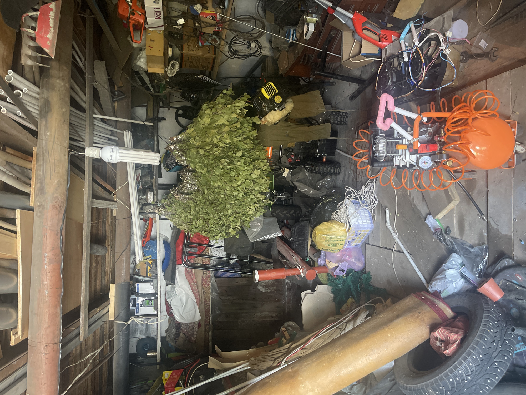 The image depicts a cluttered storage area, possibly a garage or workshop, located in the city of Sibirskiy, Russia. The space is filled with various items, including tools, construction materials, and what appear to be supplies for a bathhouse, such as birch branches.
In the foreground, an orange air compressor with a pink handle is prominently displayed. To its left, a large wooden pipe rests on a tire. The background reveals shelves stacked with boxes and equipment, as well as a collection of various tools and miscellaneous items. The overall impression is one of disorganization and storage of an eclectic mix of goods. The lighting suggests it is daytime, though artificial lighting is also visible. No people are present in the image.