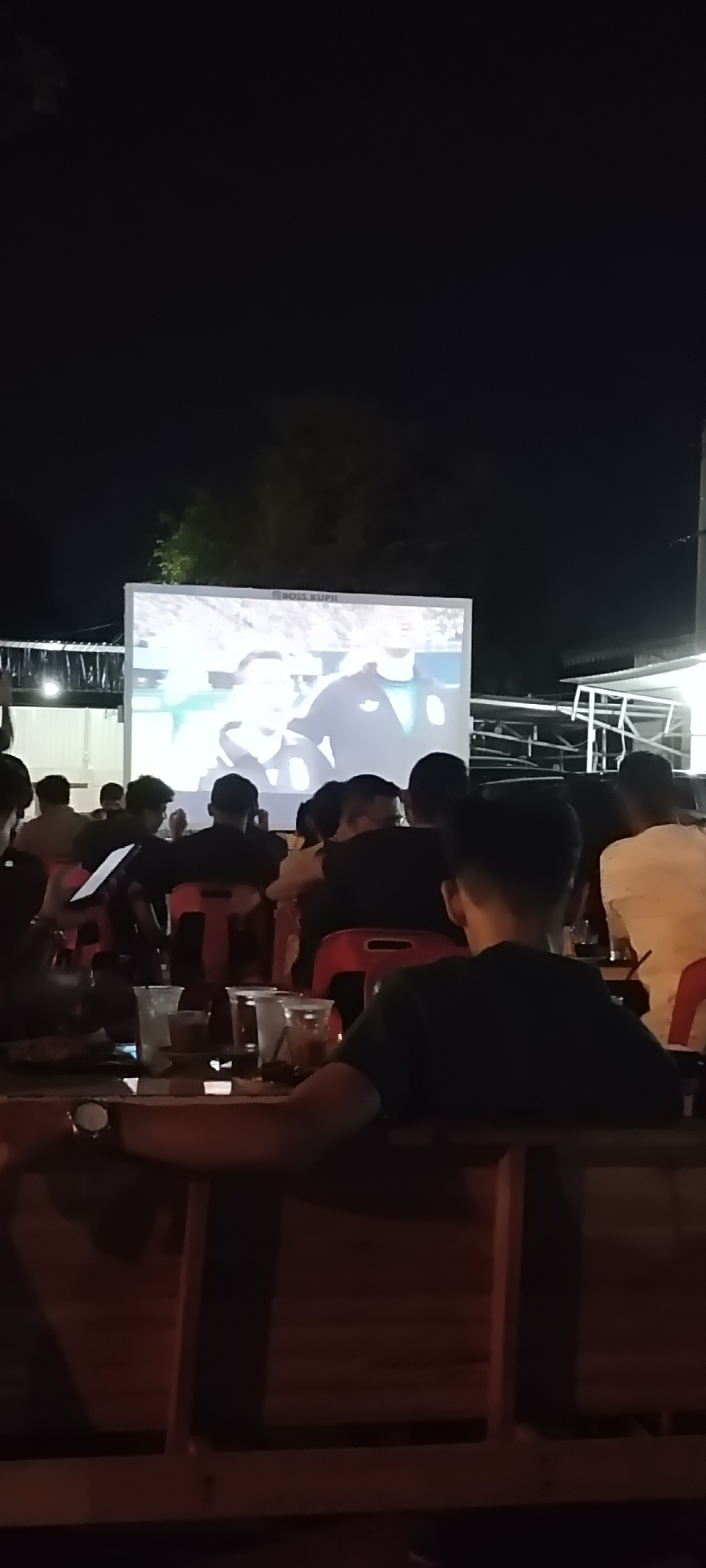 An outdoor screening is taking place, likely at a cafe or public gathering space in Banda Aceh, Indonesia, as suggested by the atmosphere and attire. A large projector screen displays a sports event or movie, with text in a logo that reads "BOSS KUPIL". A crowd of people, primarily young adults, are seated and standing, facing the screen. Many are holding drinks and some appear to be engaged with their phones. The scene is dimly lit, suggesting it is nighttime. The foreground shows tables with drinks and seating arrangements, indicating a casual, communal viewing experience. The overall impression is one of shared entertainment in a relaxed outdoor setting.