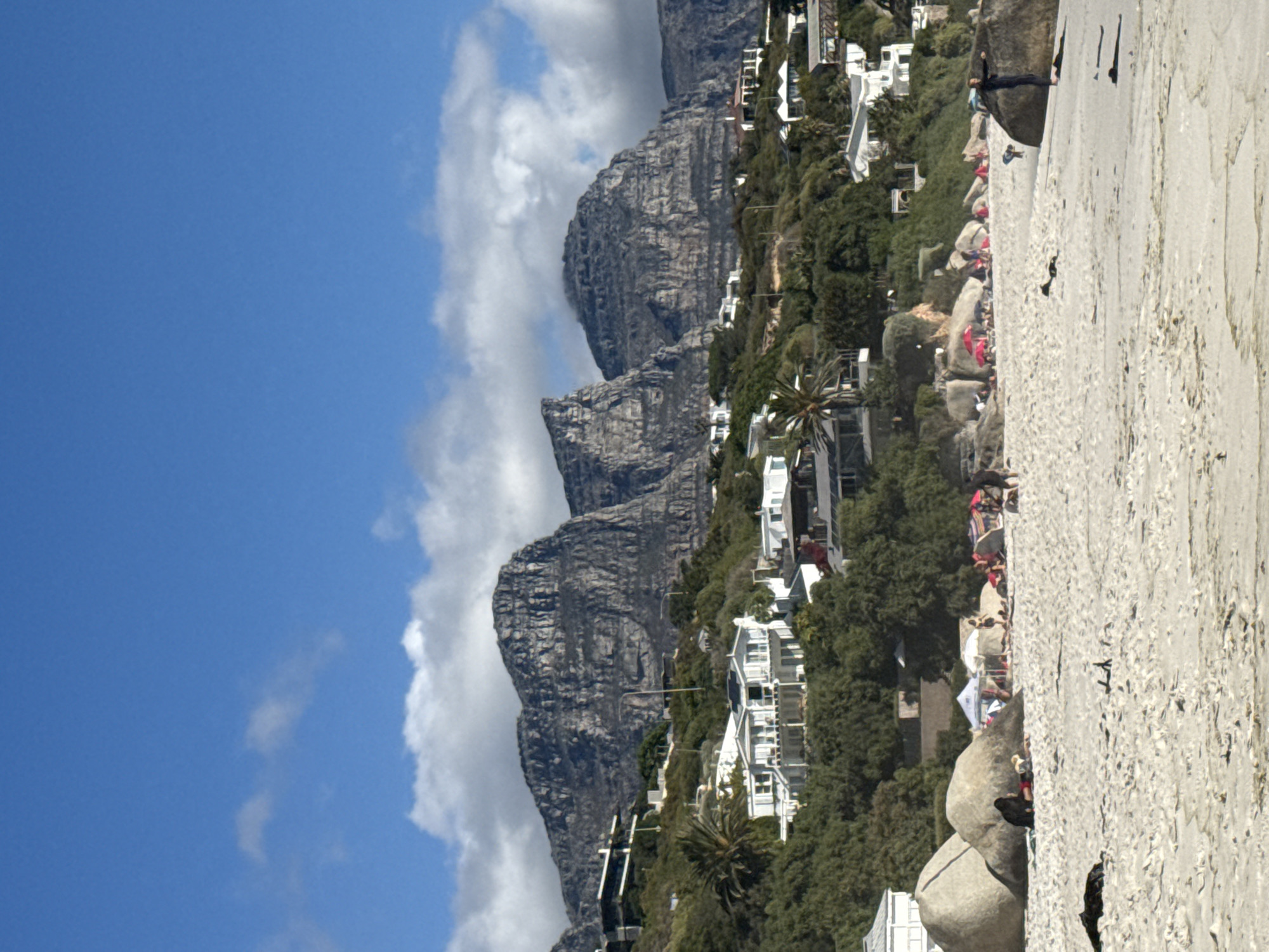The image depicts a scene at Camps Bay beach in Cape Town, South Africa, featuring a beachside view with Table Mountain in the backdrop. The foreground is dominated by a sandy beach with scattered large granite boulders. People are seen sunbathing and picnicking on the beach and around the rocks, some with beach umbrellas. Vegetation and houses line the area between the beach and the mountain, suggesting residential development along the coastline. The sky is partly cloudy with blue patches.