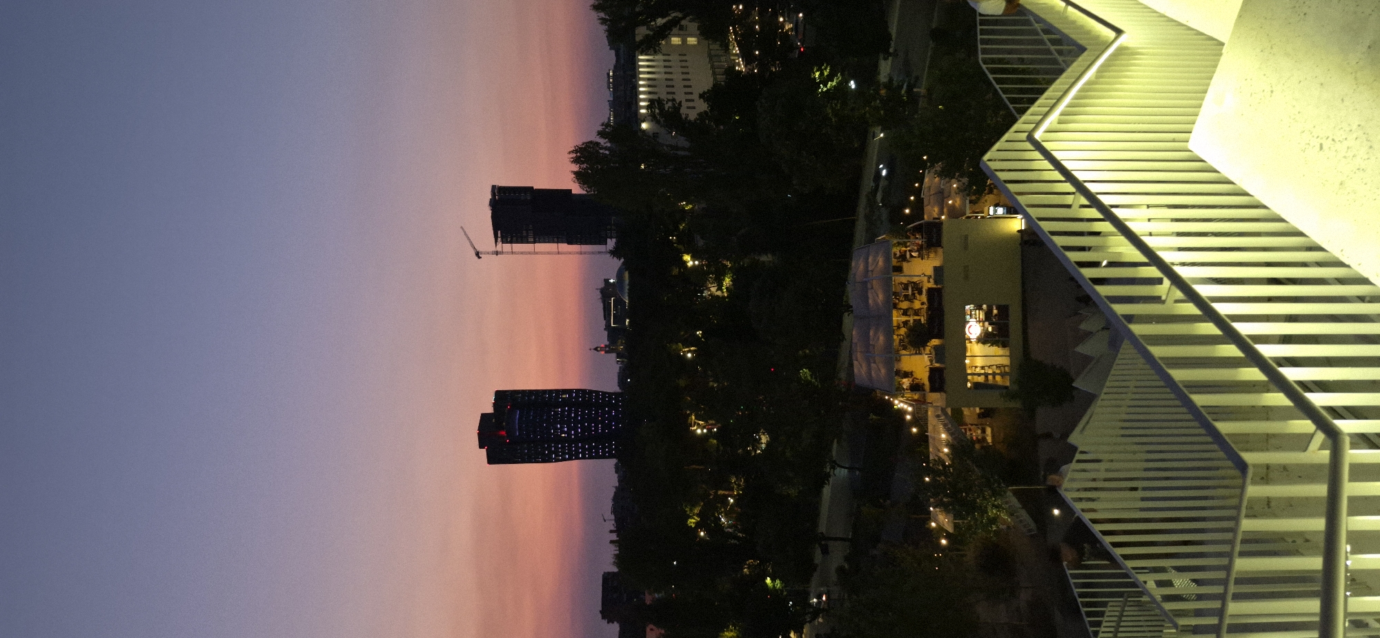 The image depicts a twilight scene in Tirana, Albania, viewed from an elevated perspective. In the foreground, a brightly lit modern staircase with white railings leads downwards, with people visible on the stairs. Below, a bustling outdoor restaurant or cafe with tables and chairs illuminated by warm lights is situated amidst lush trees. Beyond the restaurant, the city skyline features several notable buildings, including two prominent skyscrapers. One skyscraper is dark and appears to be illuminated with numerous small, bright lights, while the other is under construction, identifiable by a crane at its summit. In the background, a broader urban landscape stretches out, with more lights from buildings and streetlights dotting the scene against the fading twilight sky, which transitions from a deep blue at the top to a warm pinkish-orange hue near the horizon.