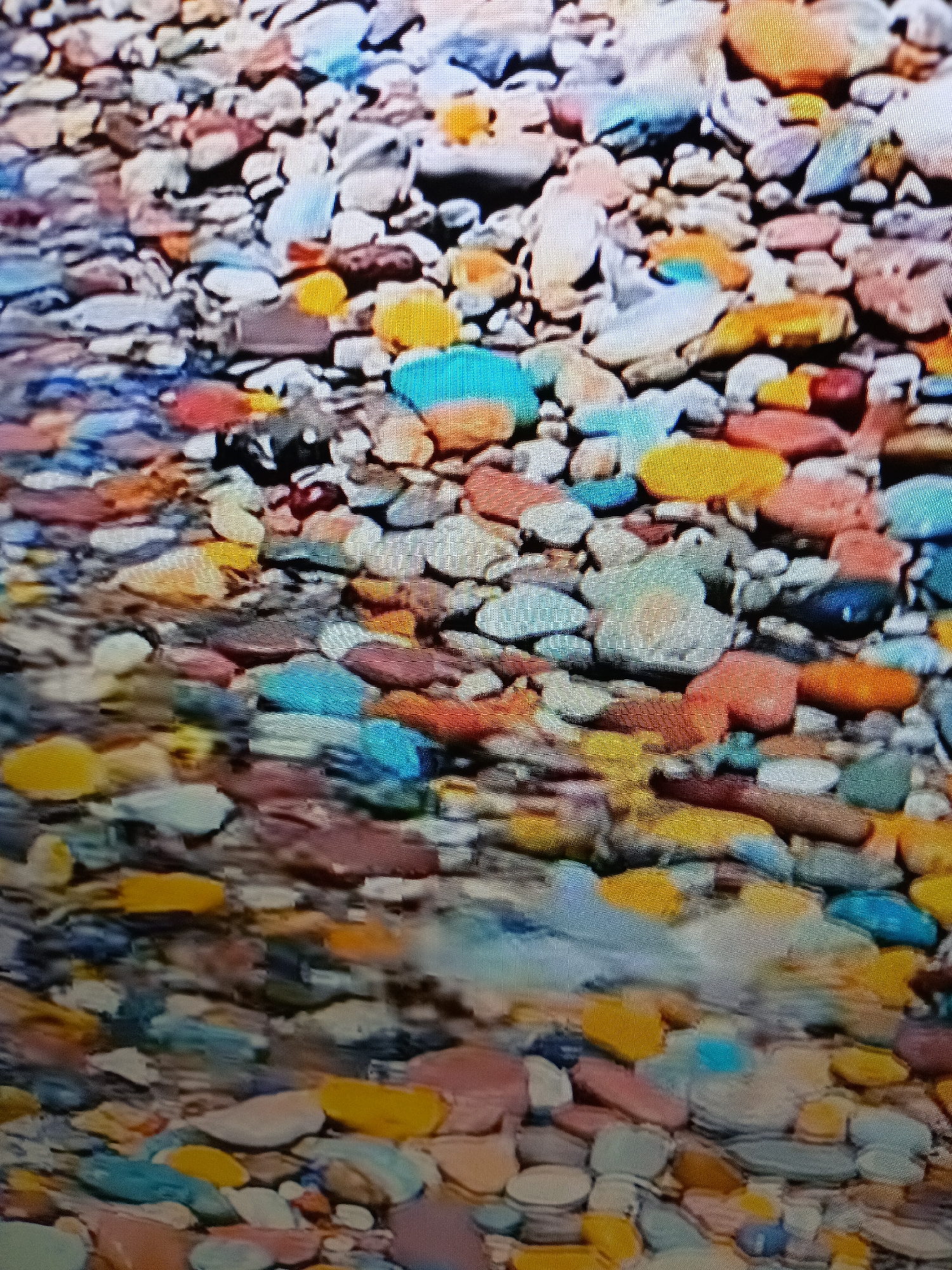 The image depicts a close-up, overhead view of numerous colorful, smooth stones submerged in shallow, clear water, likely from a riverbed or streambed. The stones vary in size, shape, and color, ranging from whites, grays, and blues to yellows, oranges, and reds, with some appearing translucent. The water creates a shimmering effect, distorting the view of some of the stones. The scene is natural and appears to be located in an outdoor environment. Specifically, this scene is from the Alara River in Alanya, Turkey, a popular tourist destination known for its natural beauty and historical sites such as Alara Castle.
