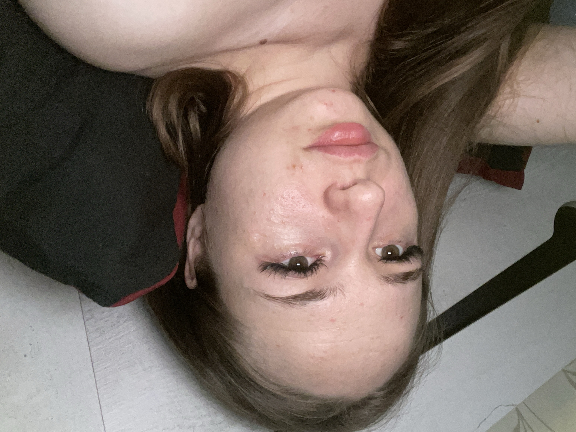 An overhead, close-up, horizontal, slightly overexposed shot shows the face of a young woman with brown hair. She is lying down, with her head tilted back and her face angled towards the camera. Her eyes are open and looking upwards. Her lips are slightly parted and appear glossy. There are a few blemishes on her cheeks and forehead. Her eyelashes are long and dark, likely enhanced. The background consists of a light-colored floor or surface with faint lines, and a dark object, possibly a pillow or blanket, behind her head. A portion of her shoulder and neck are visible on the right side of the frame. The overall lighting is dim, with a single light source appearing to come from above and to the right, creating a soft shadow on her face and neck.