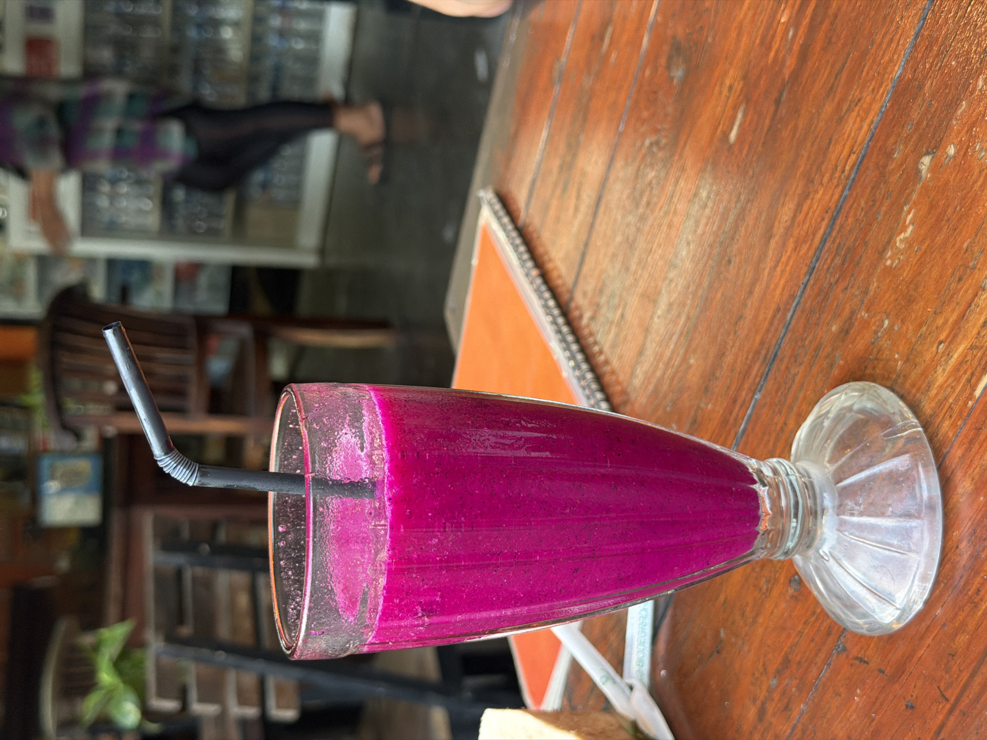 A close-up shot features a glass filled with a vibrant pink dragon fruit smoothie on a wooden table. A black, corrugated plastic straw extends from the smoothie, partially obscured by the rim of the glass. The glass itself has a textured, fluted base. In the background, a person wearing a purple and green patterned shirt and dark leggings walks past a shelving unit stocked with bottles. The setting appears to be an indoor/outdoor cafe or restaurant in Kuta Selatan, Indonesia, with glimpses of wooden furniture and what might be plants in the further background. The table surface is made of rustic wooden planks.