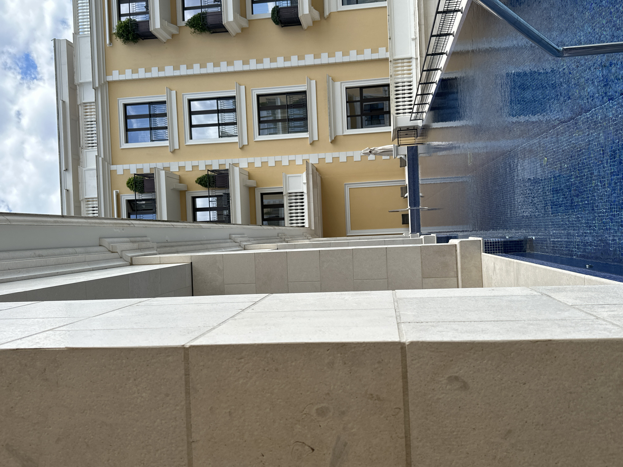 The image shows an upward view of a building adjacent to a pool, likely taken in Tivat, Montenegro. The building is several stories high, with a yellow facade and white architectural details, including window frames and decorative elements. Some windows have small planters with greenery. A metal railing is visible near the top right of the building. To the right of the building is a swimming pool with blue mosaic tiles. The foreground features beige tiled walls or steps.