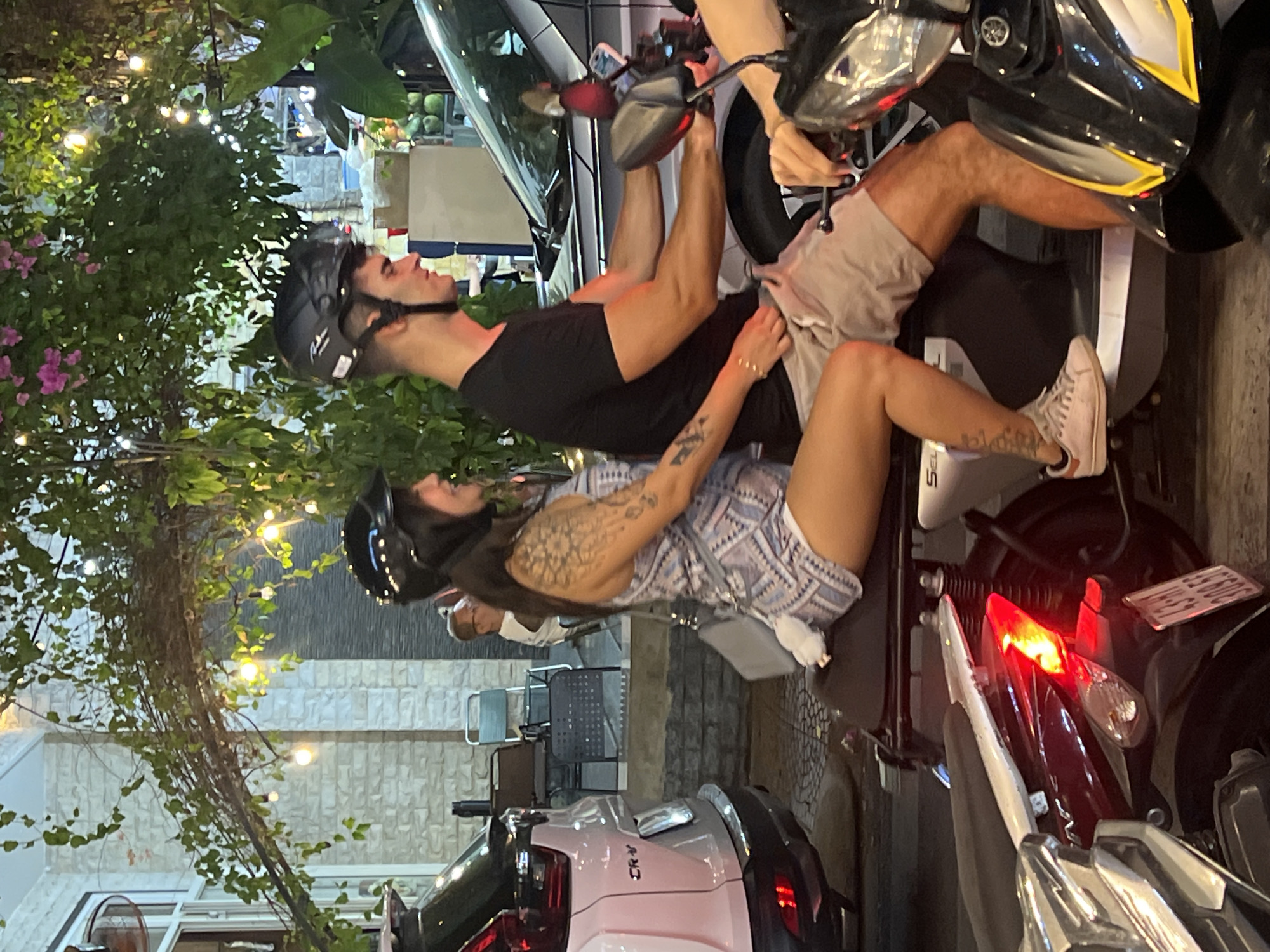 Two individuals, a male and a female, are seated on a scooter in a city street at night. The female, wearing a patterned dress and a helmet, is positioned in front. The male, also wearing a helmet and a black t-shirt, is behind her, operating the scooter. They appear to be in motion, indicated by the blur of the background and the context of riding a scooter. The environment suggests a street scene with a building in the background, foliage, and decorative lights. There are other vehicles visible, including a white SUV to the left and another scooter in the foreground. The overall scene depicts a common urban mode of transportation in Đà Nẵng, Vietnam, where scooters are widely used.