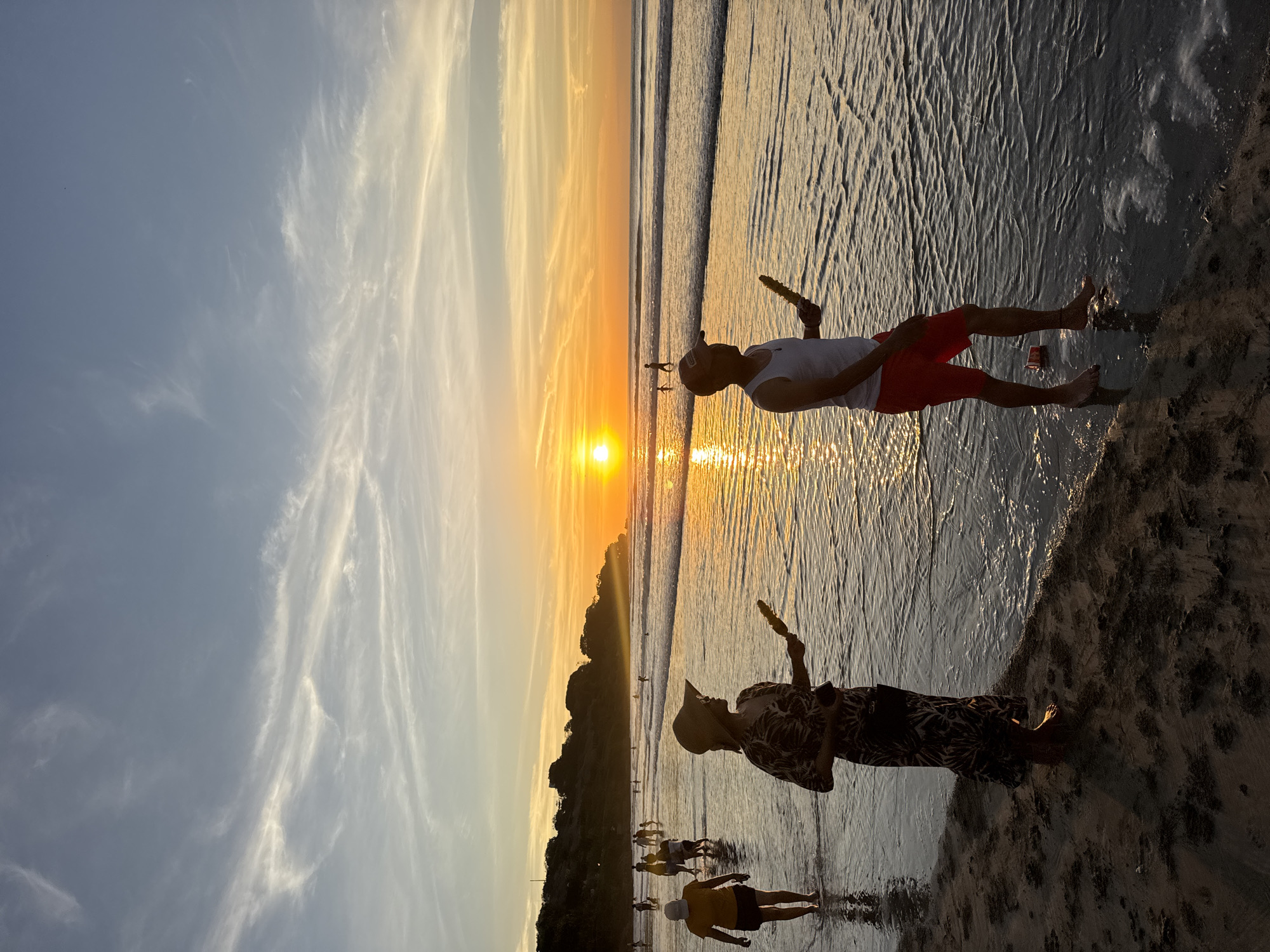 The image depicts a beach scene at sunset in Kuta Selatan, Indonesia. The sun is setting over the ocean, casting a golden glow on the water and sky. Two people are standing in the foreground. The person on the right is wearing red shorts and a white top. The person on the left is wearing a patterned outfit and a hat. Both are holding objects. Several other people are visible in the distance along the shoreline. The background includes a forested area. The water is calm, and the sky has streaks of clouds.