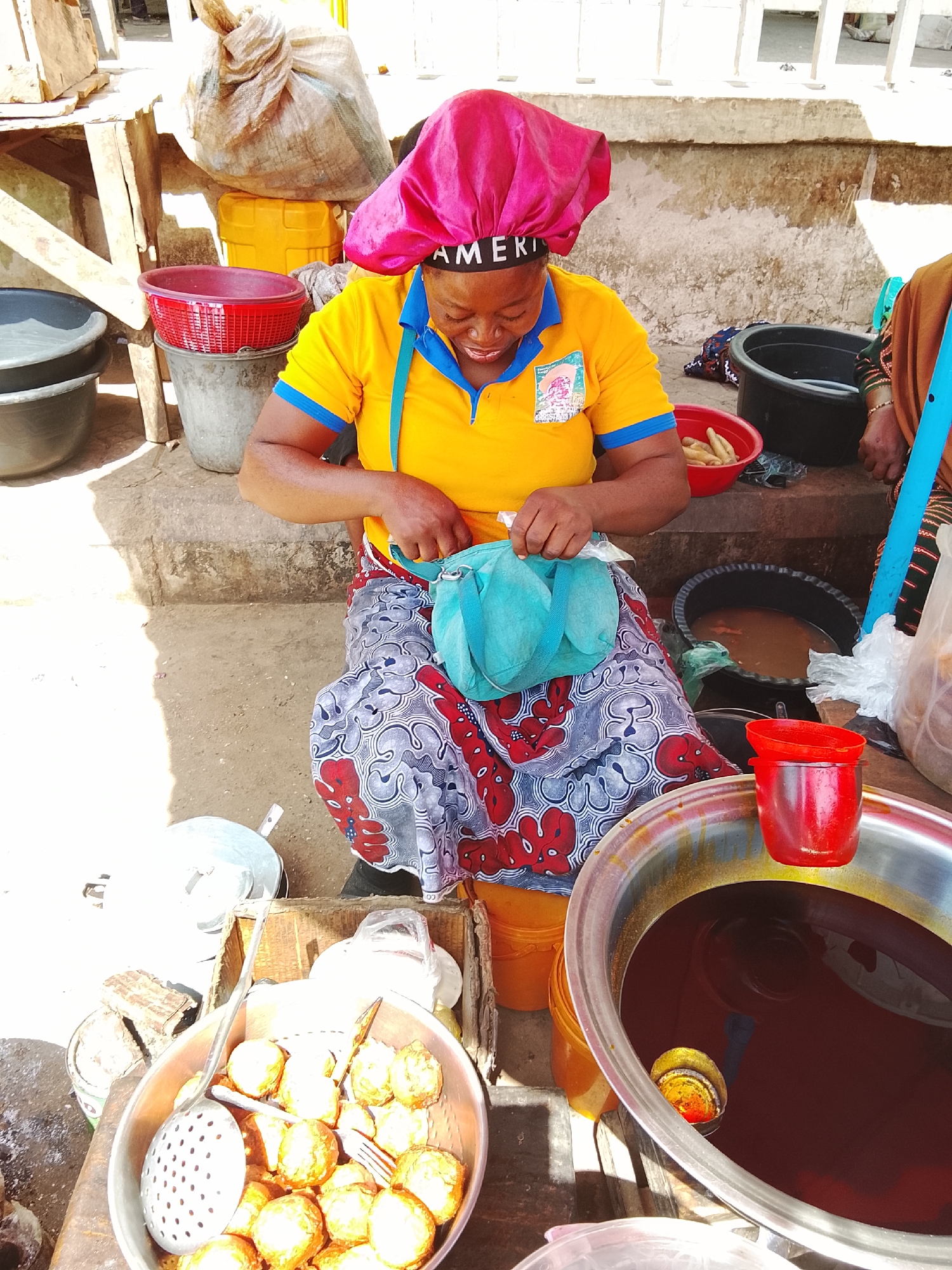 An adult female is centrally positioned, seated on a raised concrete step at an outdoor food stall in Akwanga, Nigeria. She wears a yellow polo shirt with blue collar and sleeve trim, a red, blue, and white patterned skirt, and a bright pink satin head covering with "AMERICA" printed in black on its band. Her head is bowed, and her hands are actively manipulating a light blue fabric item, possibly a pouch, held over her lap.
To the subject's left foreground, a metal bowl contains approximately two dozen spherical, golden-brown fried food items, alongside a metal slotted spoon and a two-pronged fork. To her right foreground, a large, shallow metal basin holds a dark red liquid, consistent with palm oil, in which a yellow and red ladle is partially submerged. A red plastic funnel-shaped object rests on the basin's rim.
On the concrete step behind the subject, a red plastic bowl contains several peeled, elongated tubers, and a black plastic basin holds a dark, opaque liquid. The immediate ground level consists of light-colored dirt. In the background, various market items are visible, including grey and black plastic buckets, a wooden stand supporting a large beige sack, and a yellow plastic container. Partial views of concrete structures and railings are also present in the background.