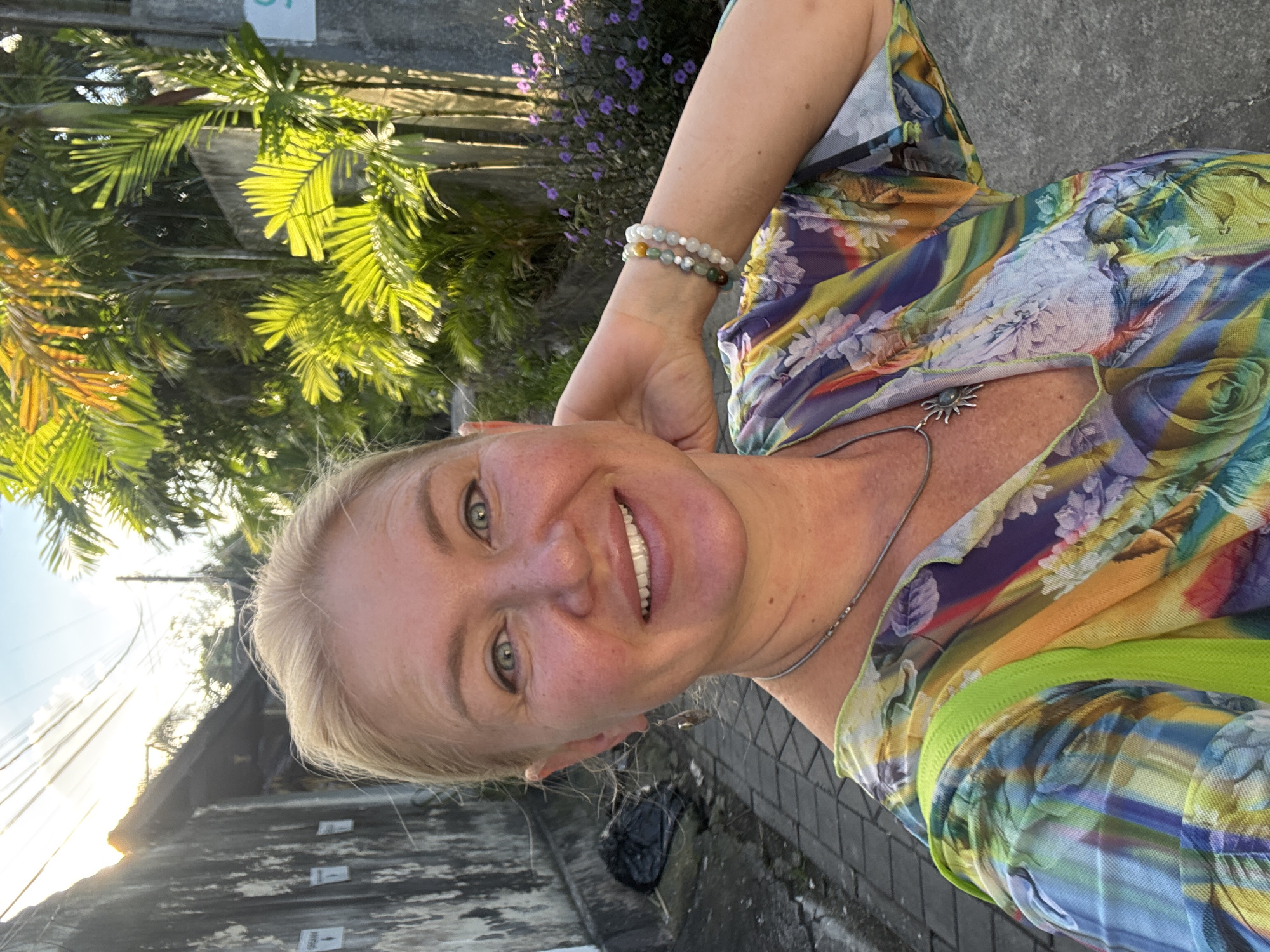 A light-skinned woman with blonde hair smiles at the camera in an outdoor setting. She wears a colorful, floral-patterned top, bracelets, and a necklace with a sun pendant. Lush green palm trees and purple flowers grow behind her. The location appears to be a street in Kuta, Indonesia, evident from the tropical foliage and architectural styles. The street is paved with grey bricks, and there's a dark object resembling a trash bag nearby. Sunlight is strong, suggesting a daytime setting.