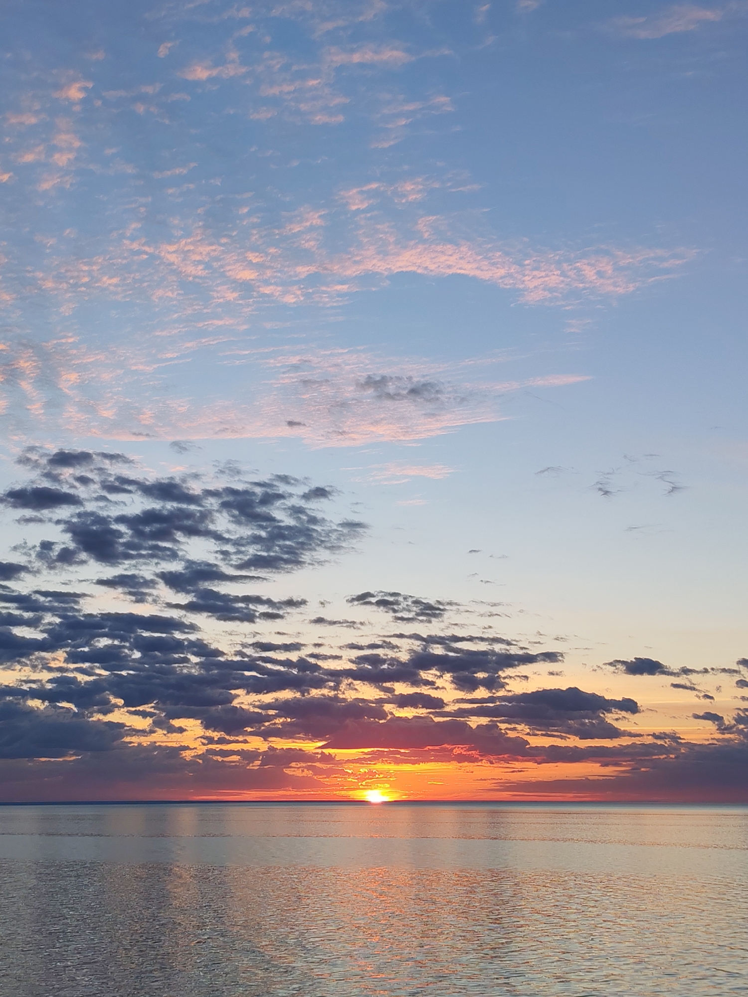 The image captures a sunset over a body of water, likely near Bol'shie Mkhi, Russia, given the location context. The sun is positioned low on the horizon, partially obscured by a layer of dark, scattered clouds. The sky transitions from light blue at the top, interspersed with thin, pink-tinged clouds, to orange and red closer to the horizon where the sun is setting. The water reflects the colors of the sunset, with shimmering orange and pink hues. The water appears calm with small ripples across its surface.