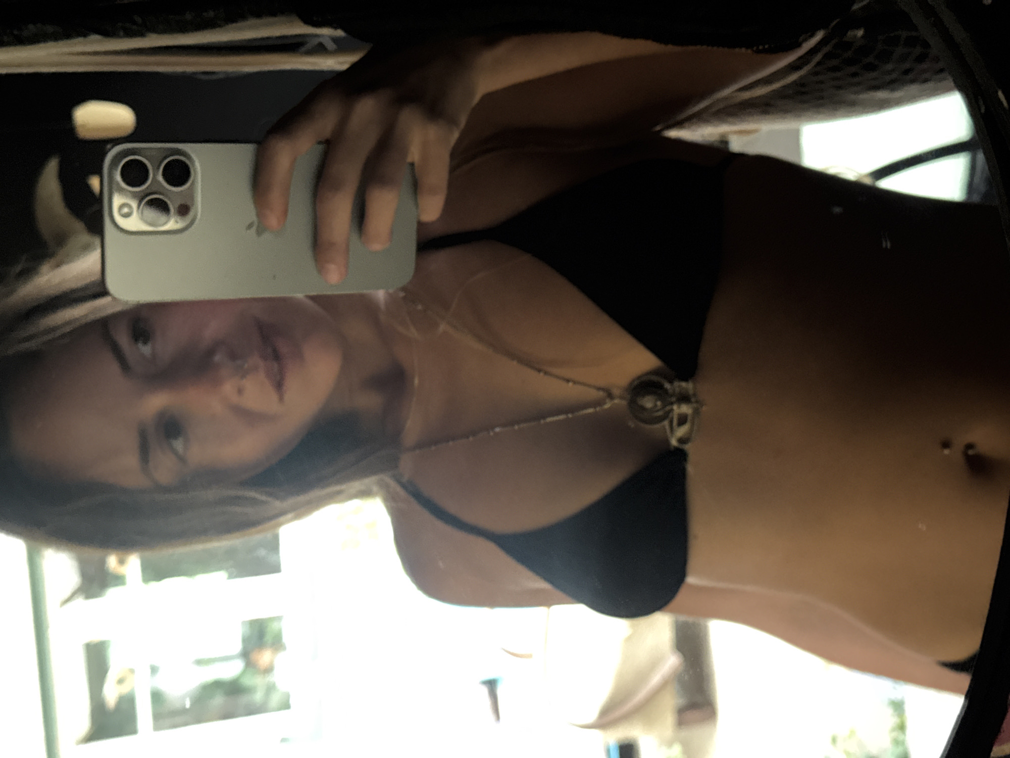 In a mirror selfie, a woman with long, dark hair and a nose ring stands in what appears to be a dimly lit room with natural light filtering in from a window. She is wearing a black bikini top and a necklace with a decorative pendant. She is holding a light green smartphone in her right hand, angled to capture her reflection. The background is blurred, but hints of greenery and possibly furniture can be seen through a window or opening. The woman's face is partially obscured by her hair and the phone.