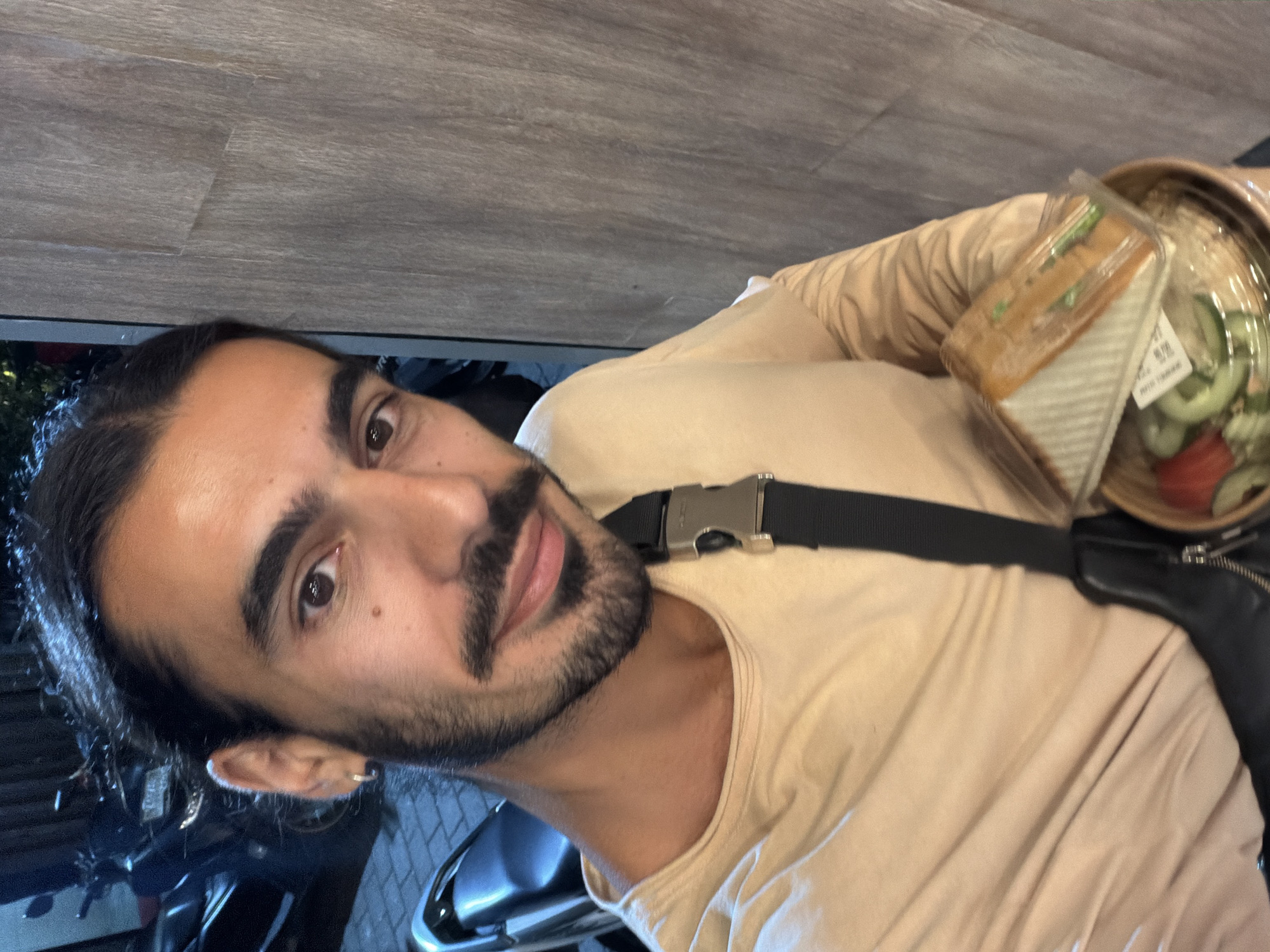 The image is a vertical, close-up selfie of a man taken at night or in the evening. The man has dark, long hair tied back, a prominent mustache, and a short beard. He wears a light beige or tan short-sleeved t-shirt. A black strap with a metallic buckle crosses his chest, part of a bag system.
He looks directly at the camera with a neutral expression. On his right side, carried alongside his body, is a black bag, partially visible with a zipper. Resting on or near this bag are two food containers. One is a clear plastic container holding a pre-packaged sandwich cut into triangles, with visible greens. A white label with unreadable black text is affixed to the sandwich packaging. The second container has a brown base and clear lid, holding a salad with visible cucumber slices and red items, likely tomatoes.
The background behind the man is dark, showing reflections of artificial light, indicating an outdoor nighttime setting. A dark, reflective surface, possibly a vehicle, is visible near his left ear. Above him, a light brown, wood-patterned surface, resembling a ceiling or overhang, is present. The scene suggests a casual outdoor setting, possibly near a food establishment in North Kuta, Indonesia.