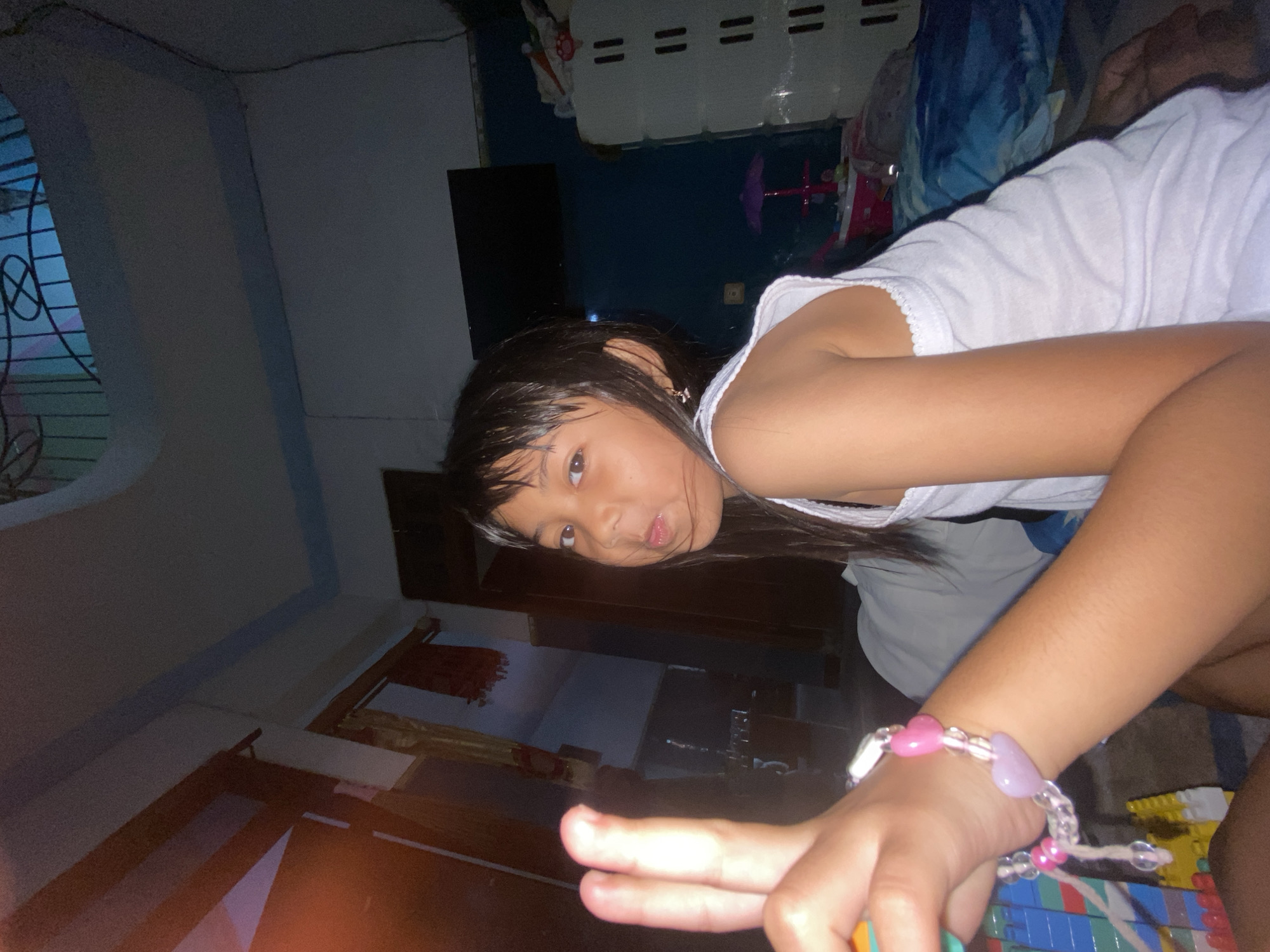 A young girl is depicted in an indoor setting, lying on her side and looking towards the camera. She has dark hair and dark eyes, and her mouth is slightly pouted. She is wearing a white sleeveless top and a pink and clear beaded bracelet on her right wrist. Her right hand is near a colorful toy, possibly building blocks.
The background shows a dimly lit room with light-colored walls, some sections featuring a blue band near the ceiling. A window with a decorative metal grille is visible on the left side of the frame. In the background, there appears to be a dark flat-screen television, a white cabinet or storage unit with horizontal dark lines, and a small purple umbrella-shaped toy. The overall lighting suggests it is either evening or night, or the room is poorly lit. The setting appears to be a residential home. No visible text is present in the image.